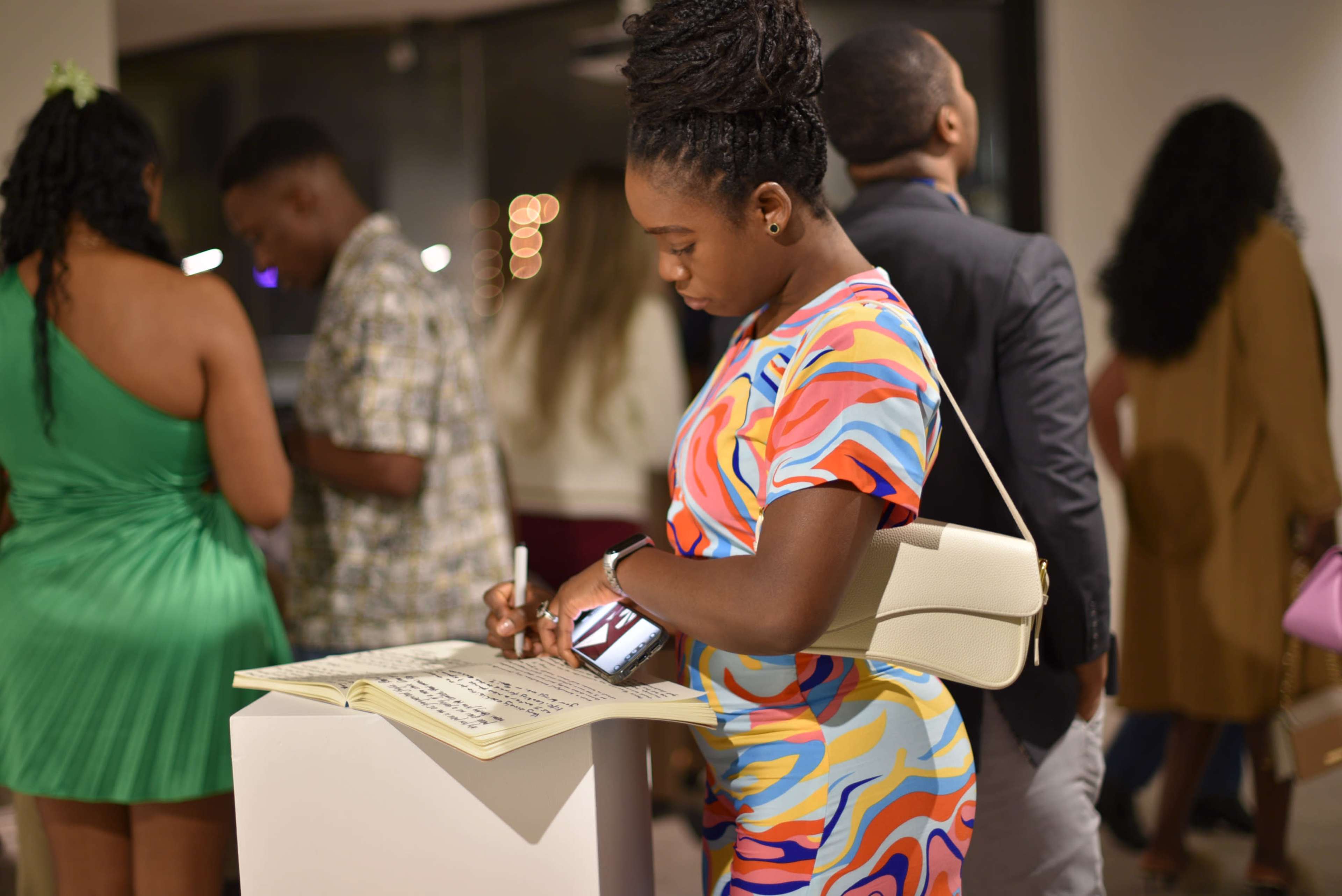 A woman in a colorful dress writes in a guestbook at an art gallery event while people mingle in the background.
