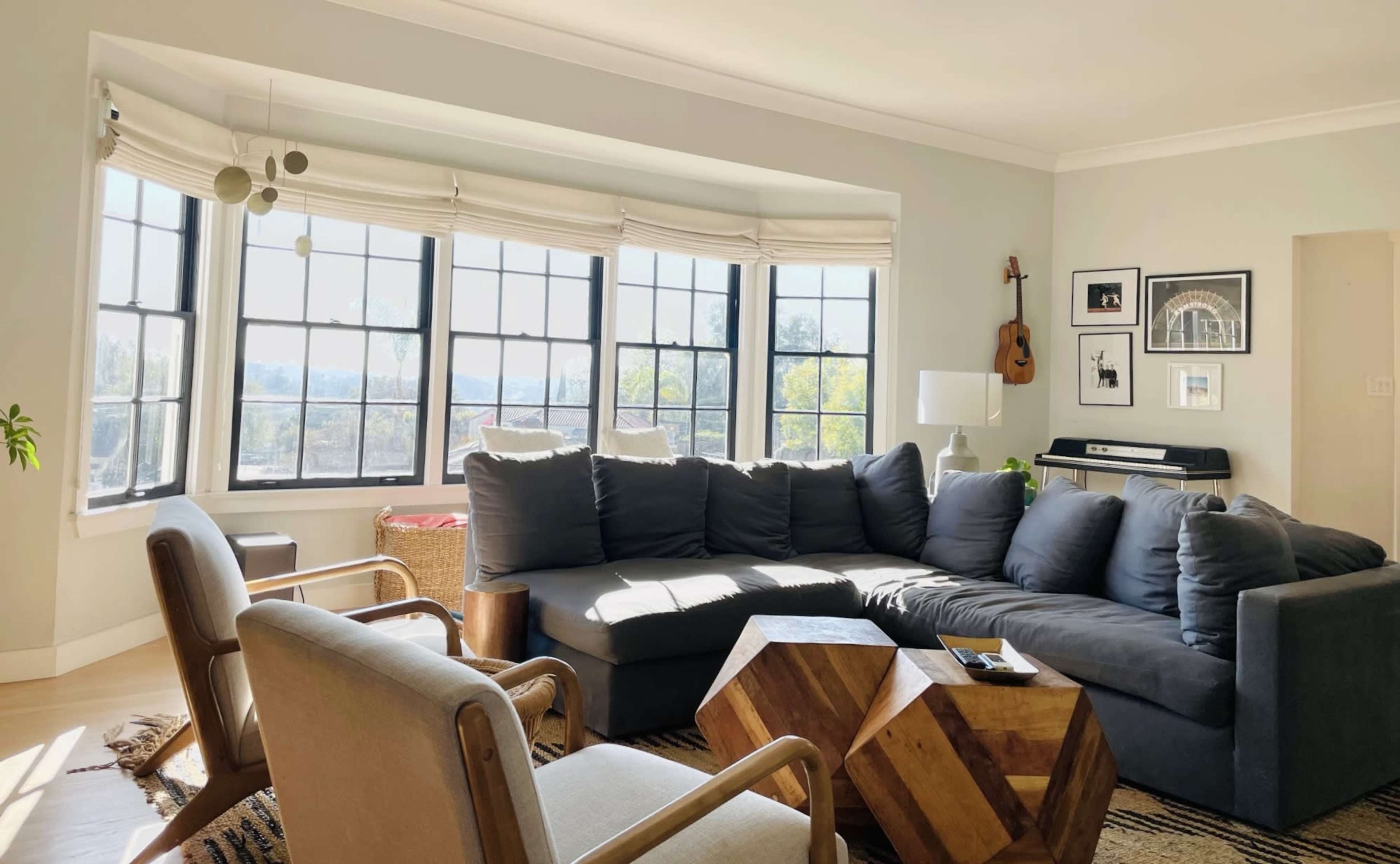 A modern living room features a large sectional sofa, an angular wooden coffee table, and windows with a view, surrounded by plants and musical instruments.