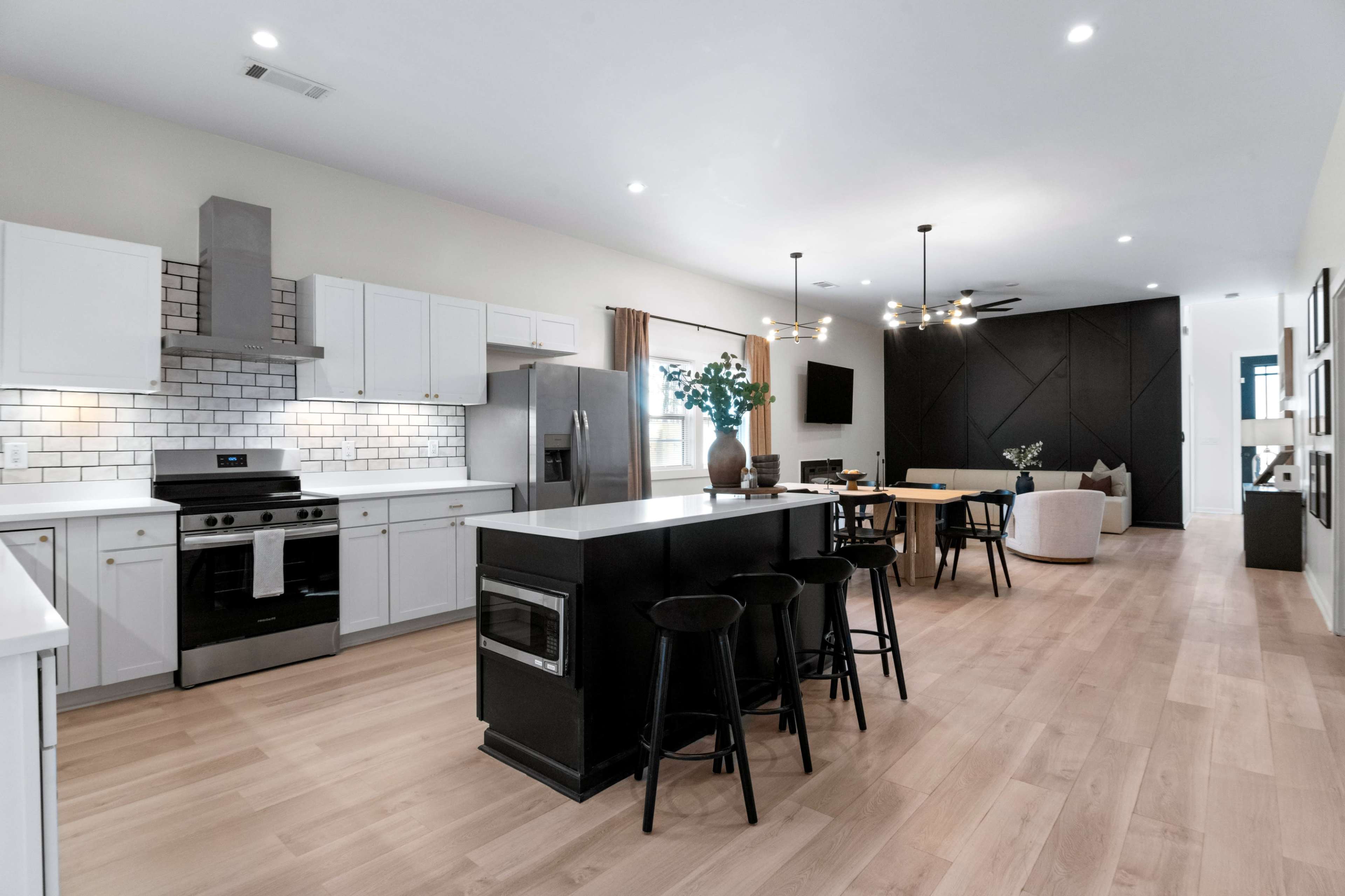 The image shows a modern kitchen and dining area with white cabinetry, a large island with black seating, and a dining table adjacent to a black accent wall.