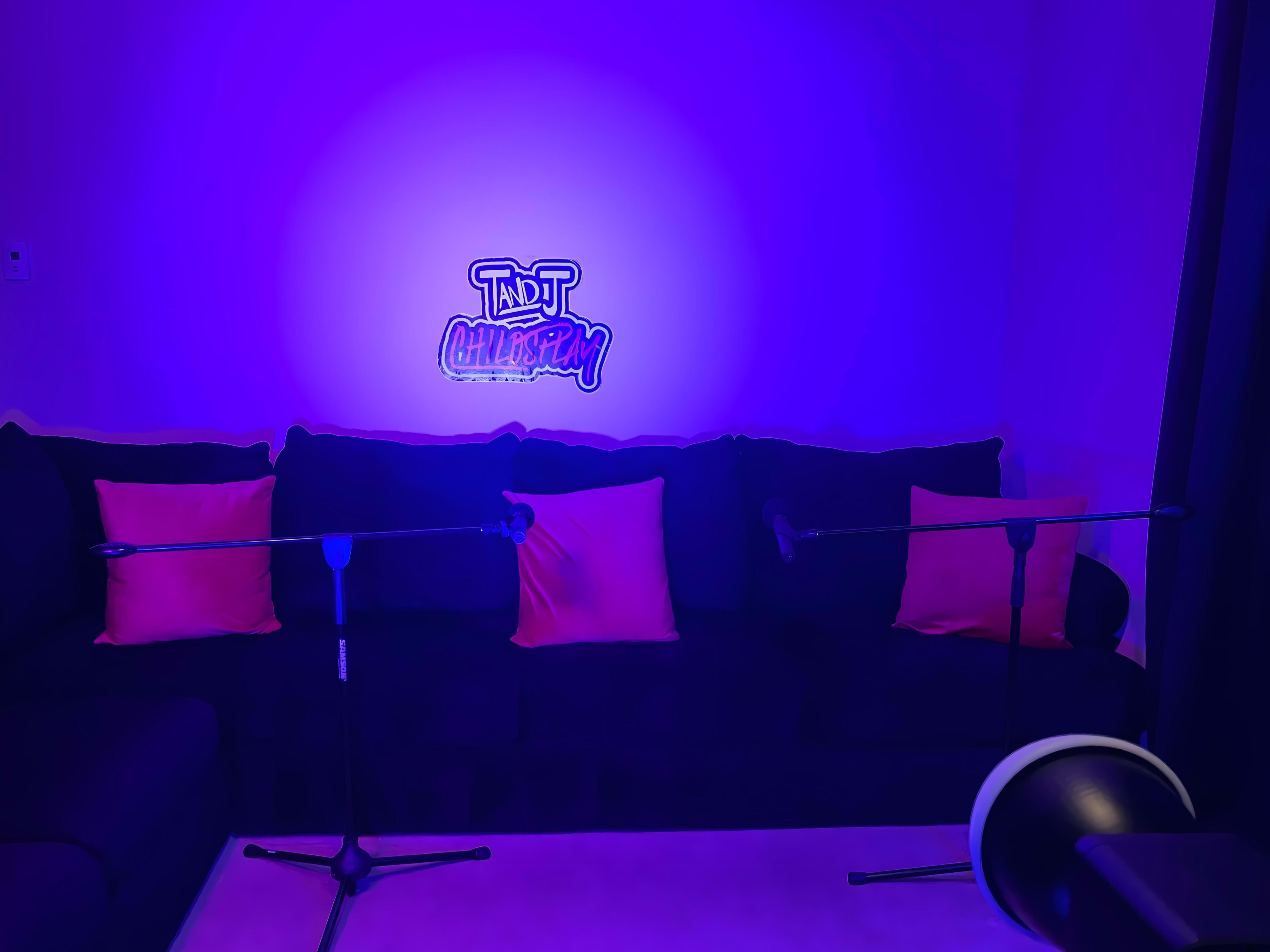 A sofa with pink cushions and two microphones is set up under a blue-purple light featuring a neon sign that reads "TNT Chats & Play."