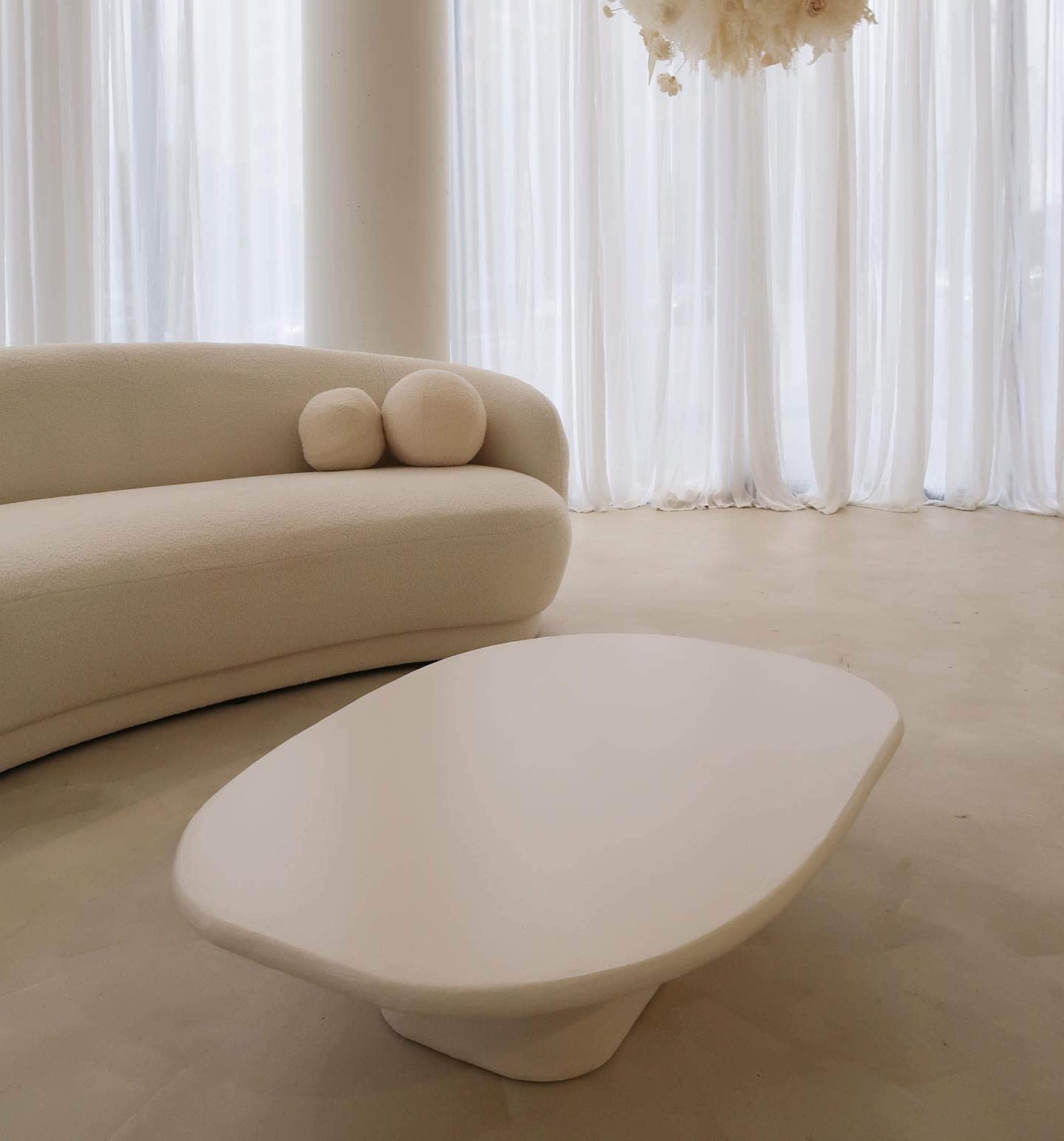 A curved, light-colored sofa with two round cushions is positioned near a sculptural, white coffee table in a bright, airy room with sheer curtains.