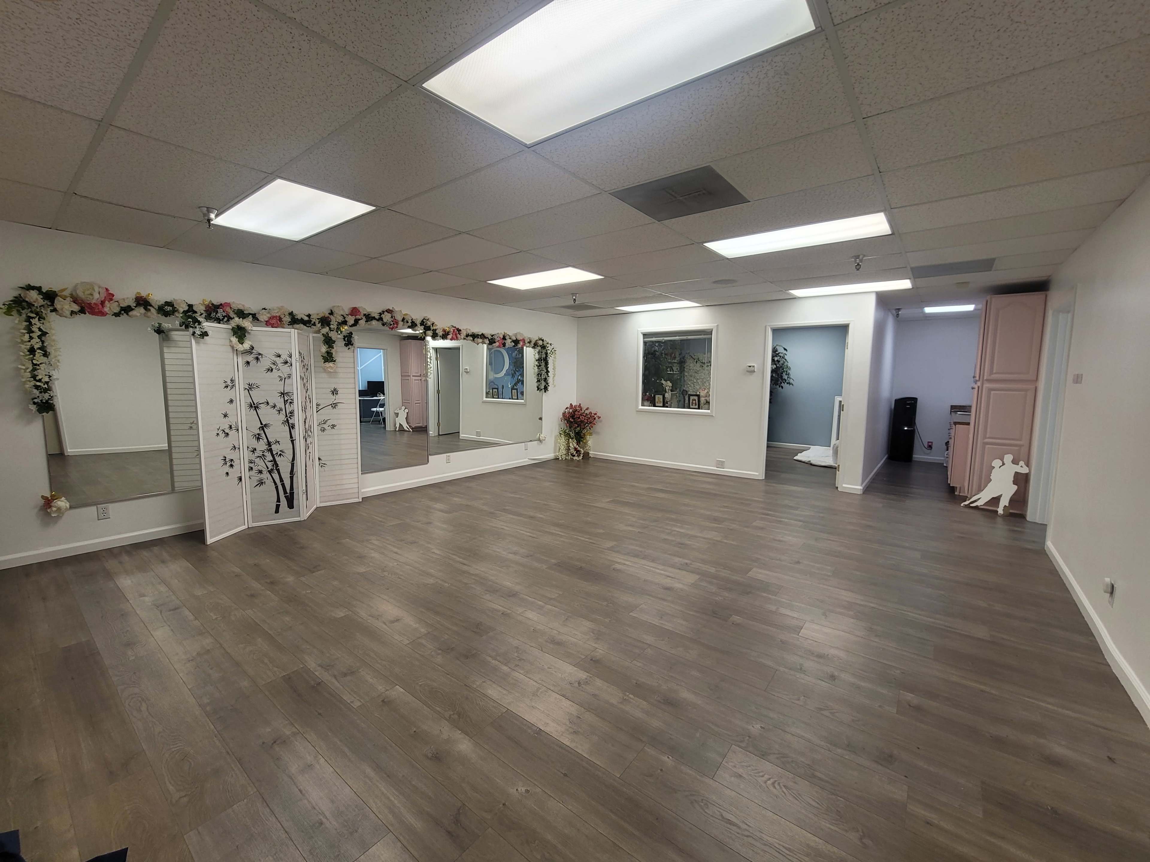 The image shows a spacious room with wooden flooring, mirrors on the walls, and decorations along the perimeter, including floral elements and a partition.