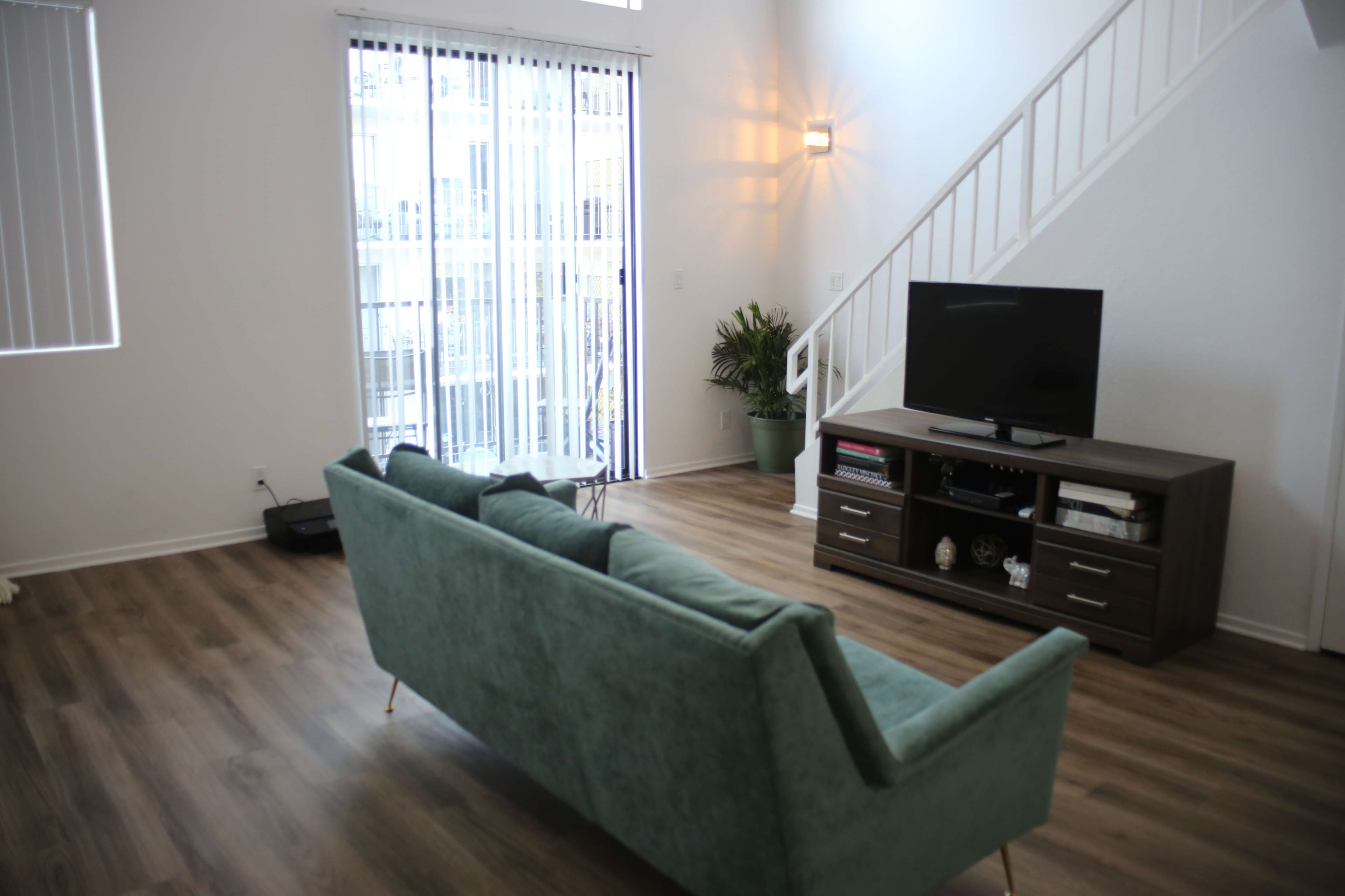 A light-filled living room features a turquoise couch facing a television set on a wooden stand, with large windows and vertical blinds.