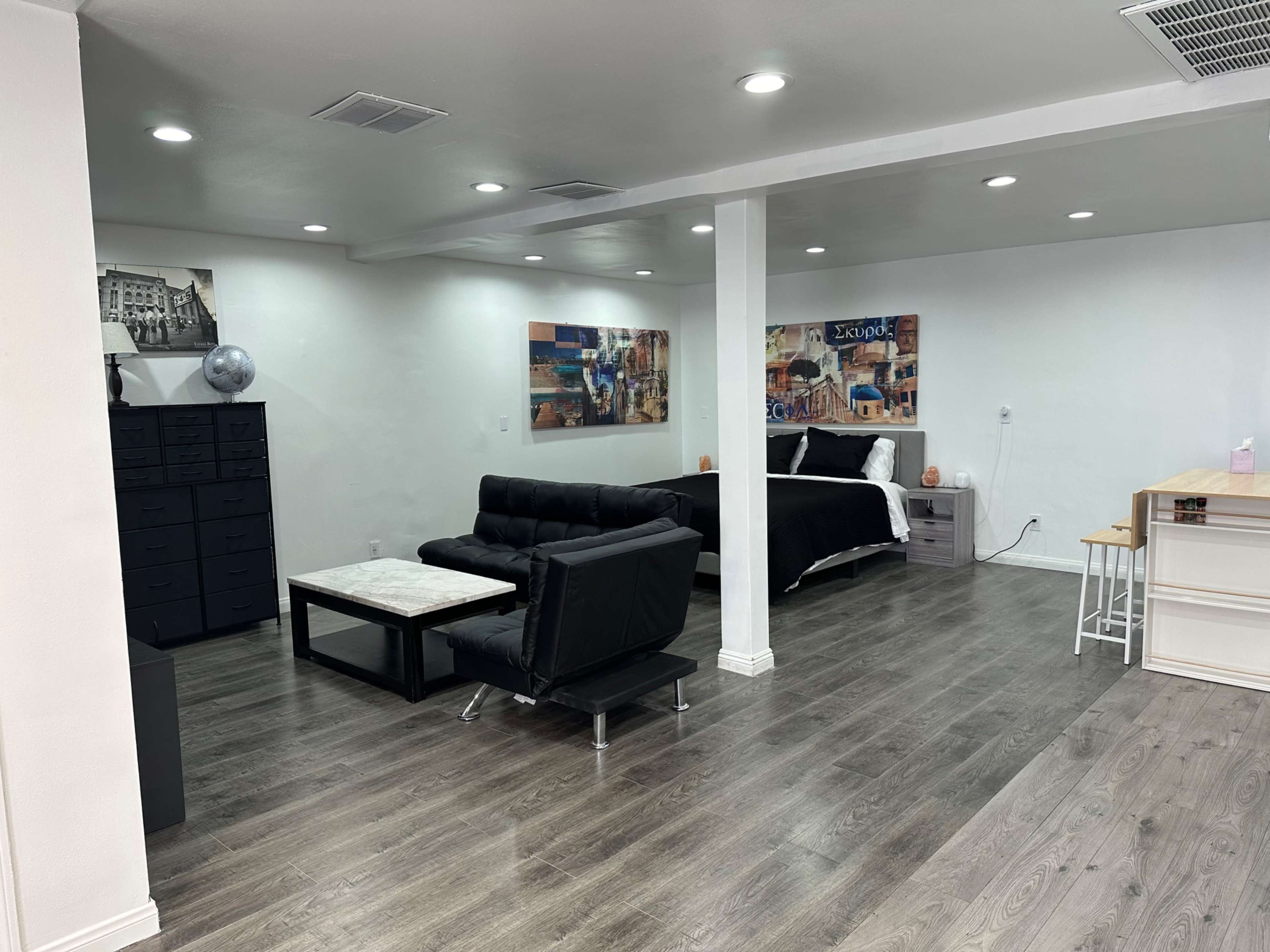 The image shows a modern, open-concept basement living area with a black sofa, a coffee table, a bed, and a kitchenette.