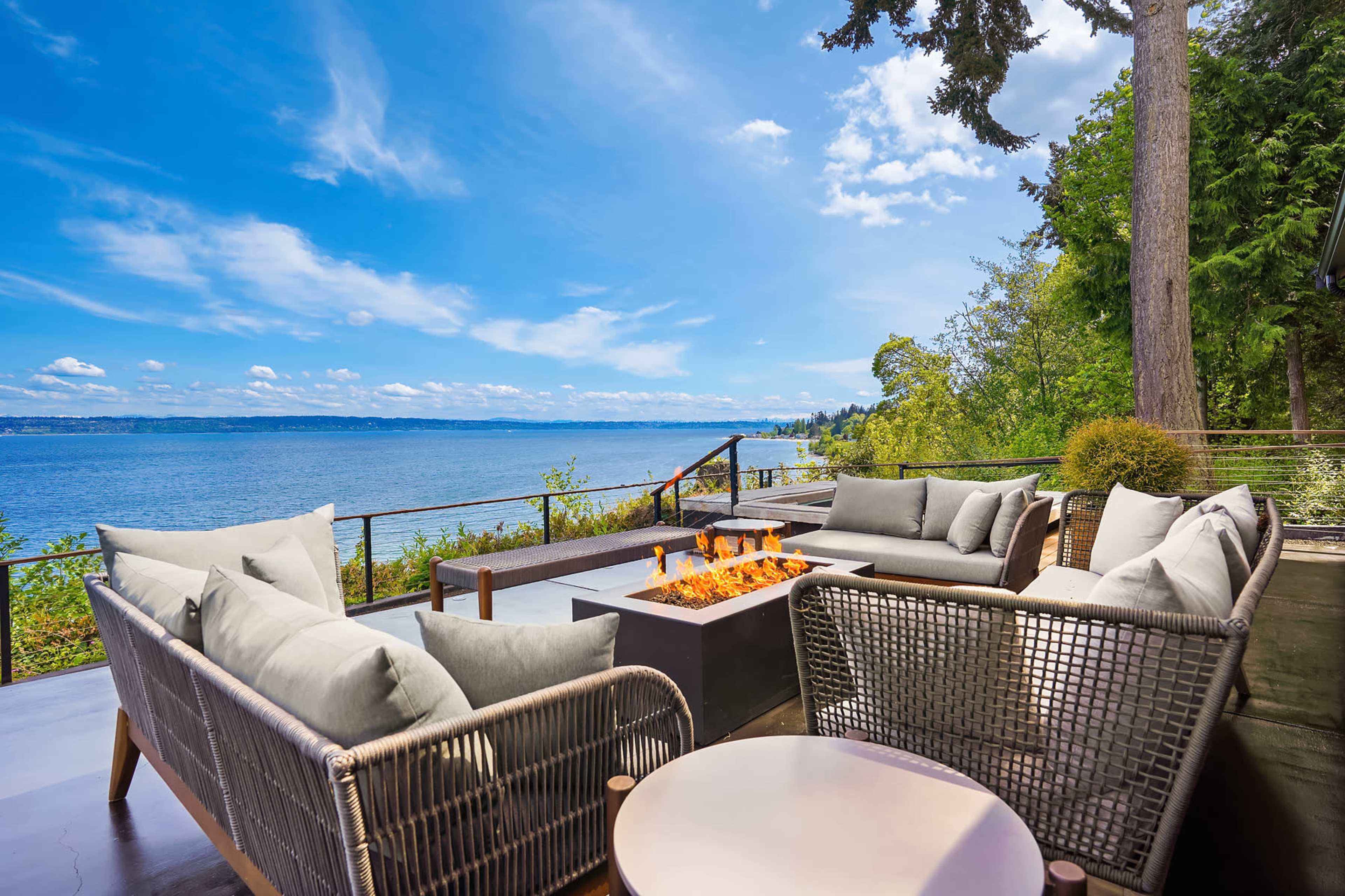 A modern patio with comfortable seating and a fire pit overlooks a calm lake under a blue sky with scattered clouds.