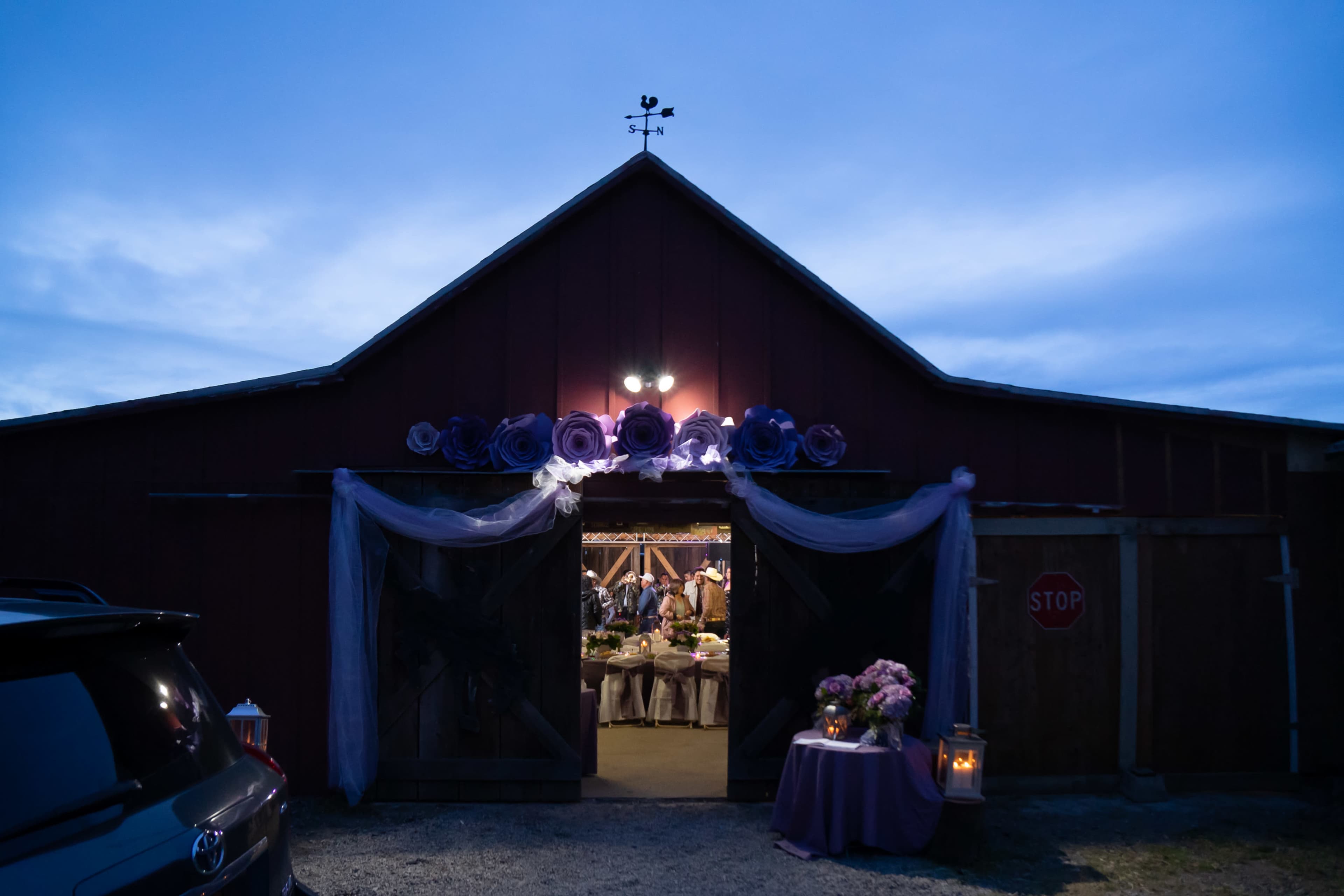 A barn with purple decorations is illuminated at dusk, revealing a festive interior and a stop sign nearby.