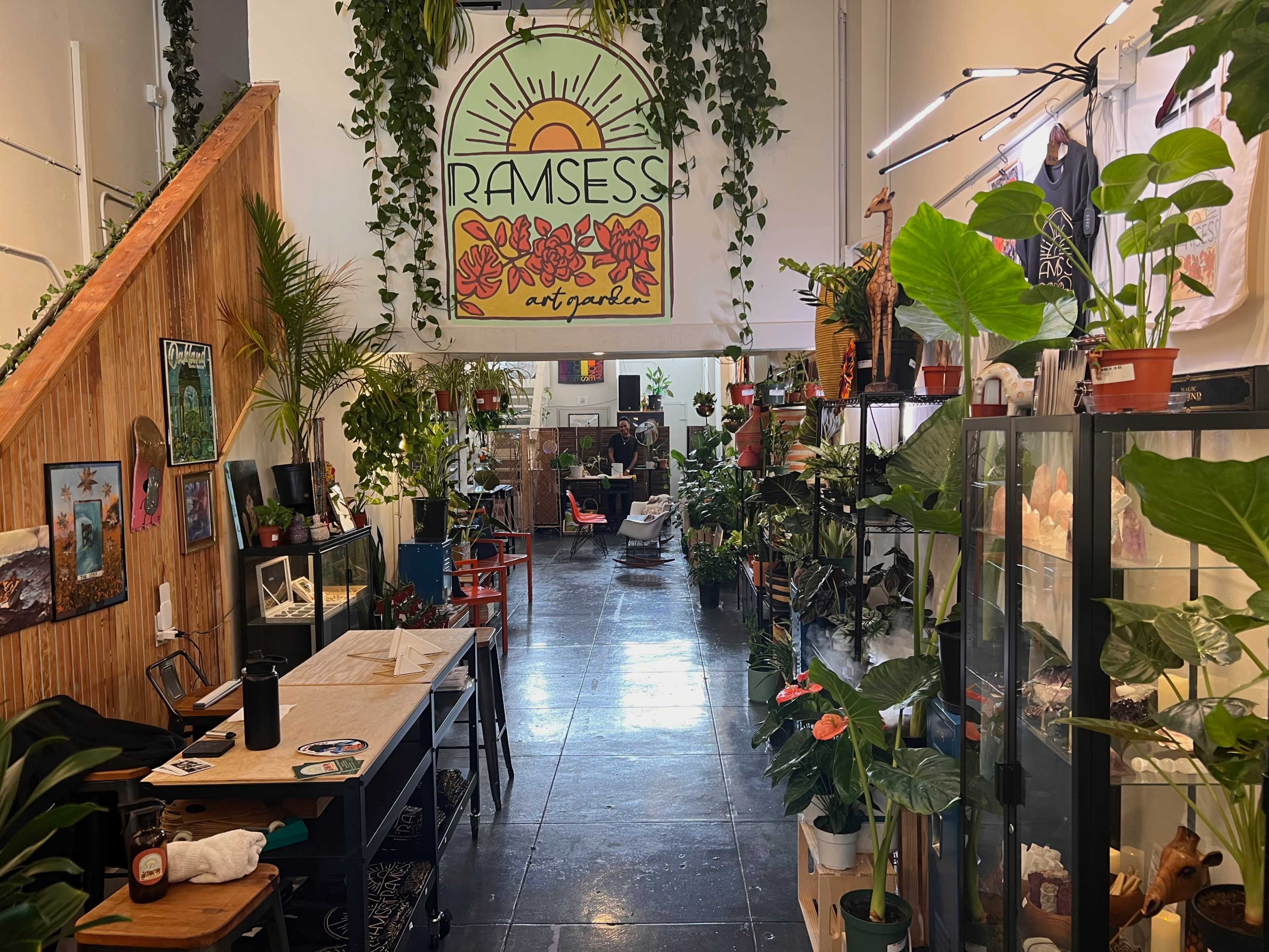 A narrow art gallery filled with plants, artwork, and seating areas, with a large sign reading "RAMSESS" hanging overhead.