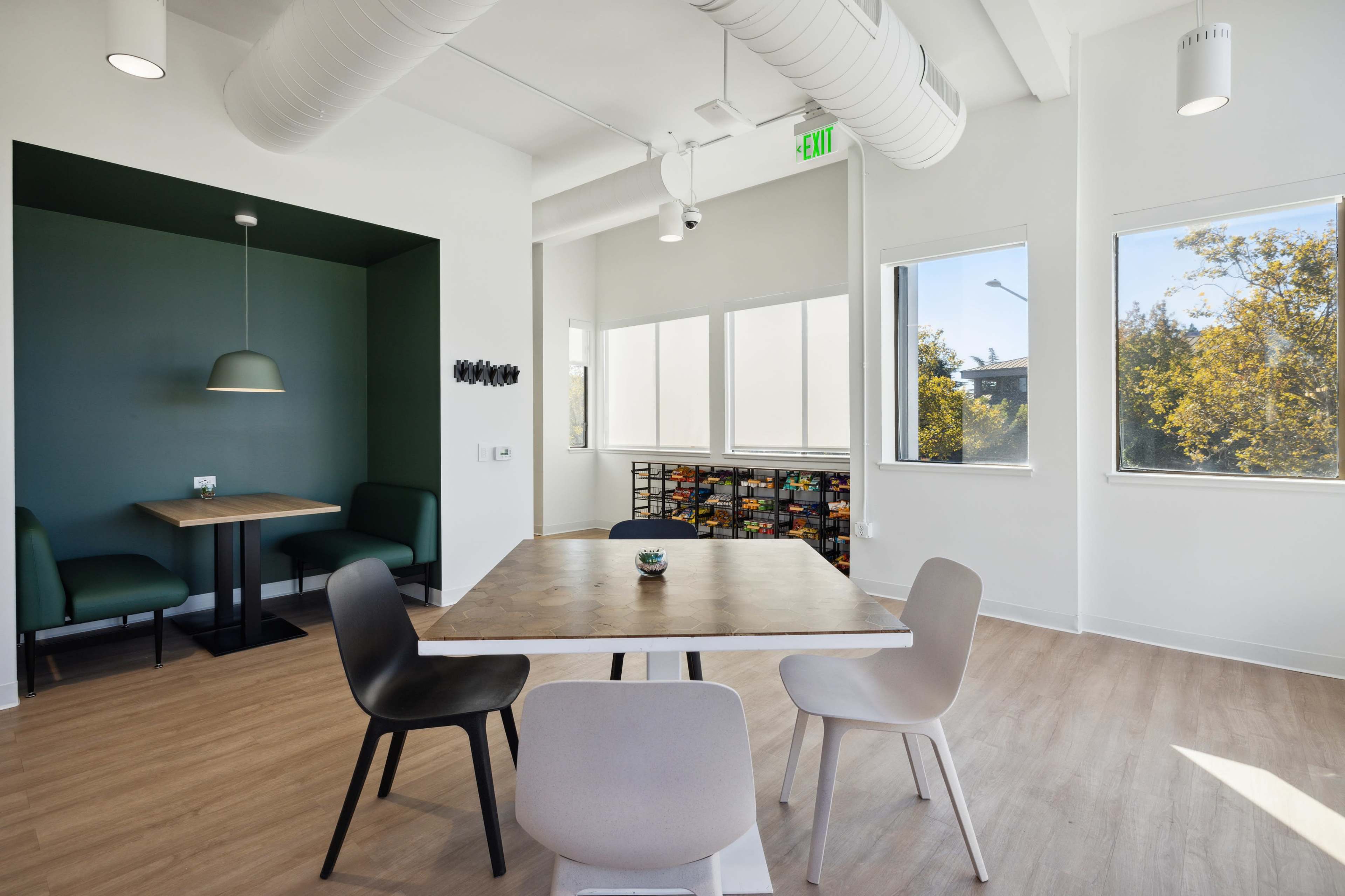 A modern open space features a wooden table surrounded by chairs, with a small dining area in the corner and large windows letting in natural light.