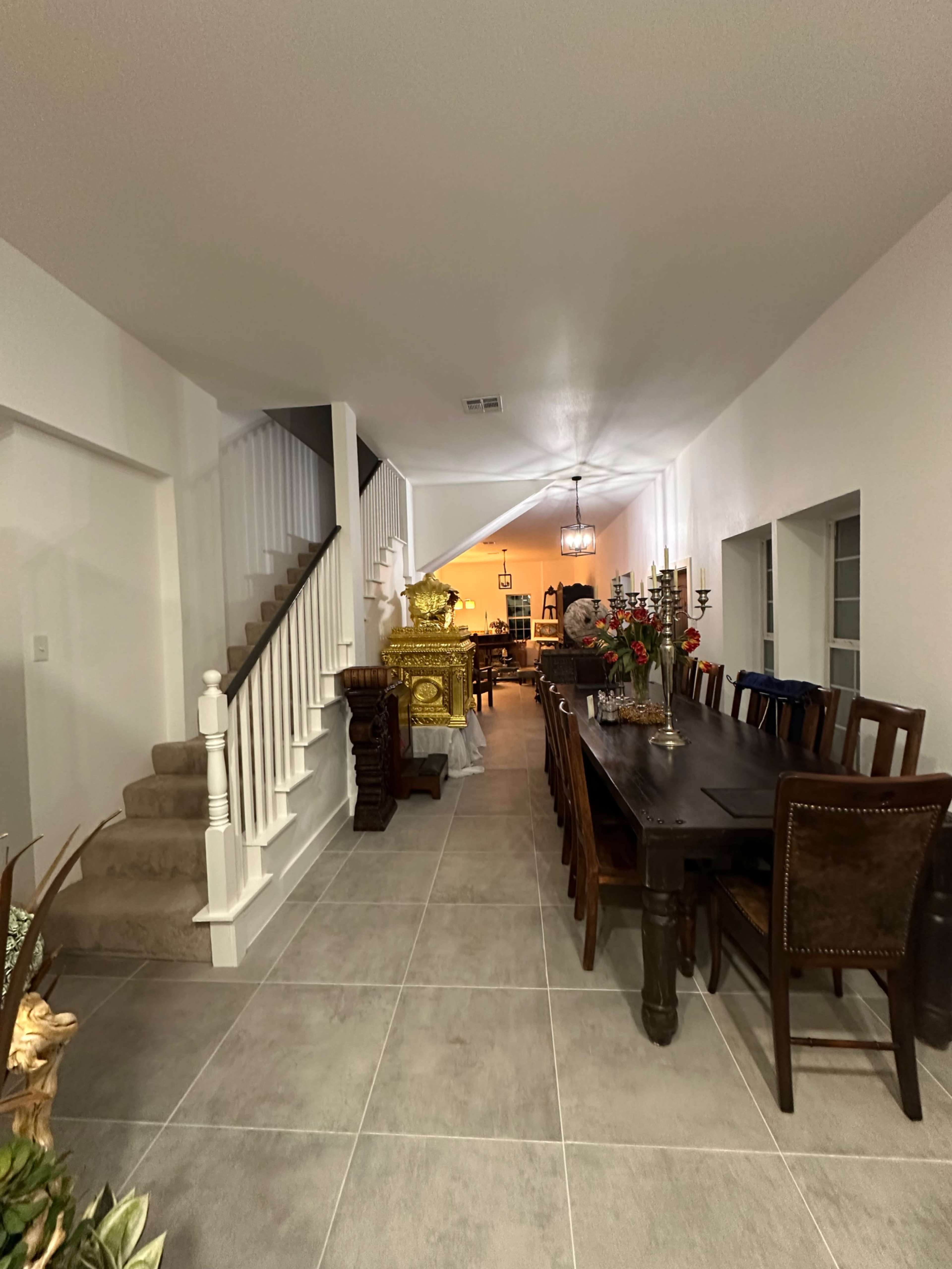 The image shows a spacious hallway leading to a dining area with a long table, stairs on the left, and decorative items in the background.