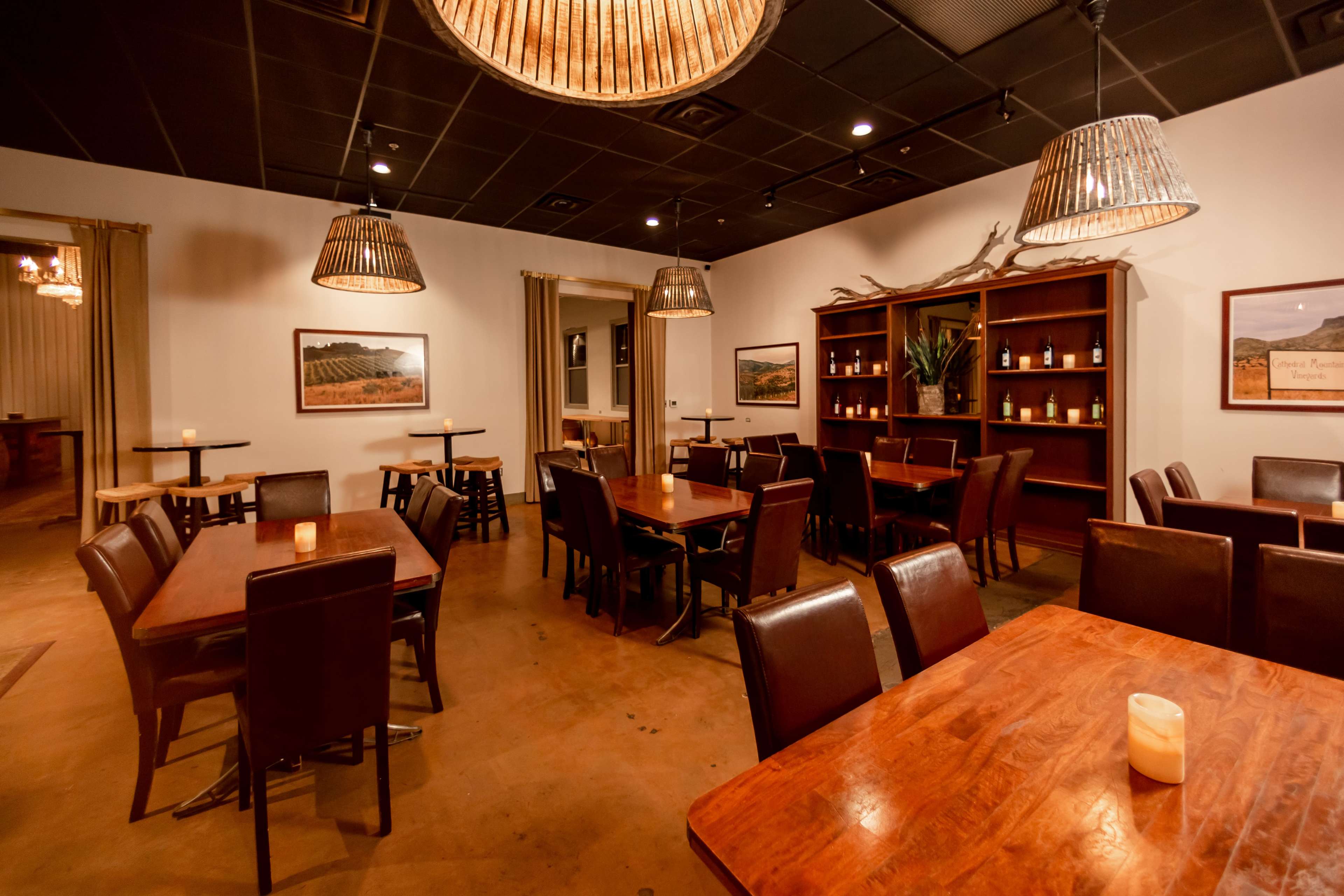 A dining area with wooden tables, chairs, and overhead lantern-style light fixtures, featuring shelves with decorative items and wine bottles.