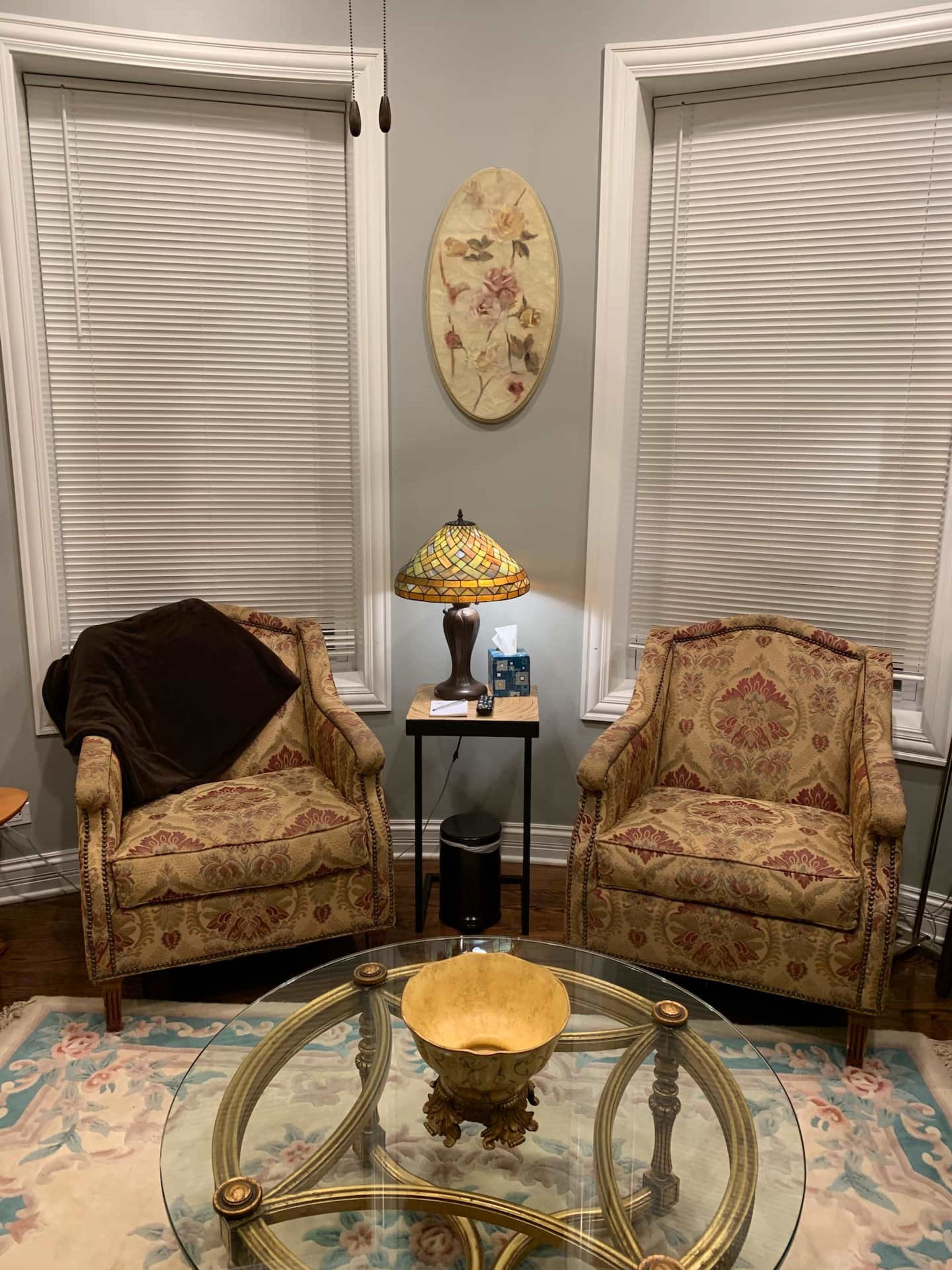 The image shows a cozy seating area with two patterned chairs, a small side table with a lamp, and a round glass coffee table.