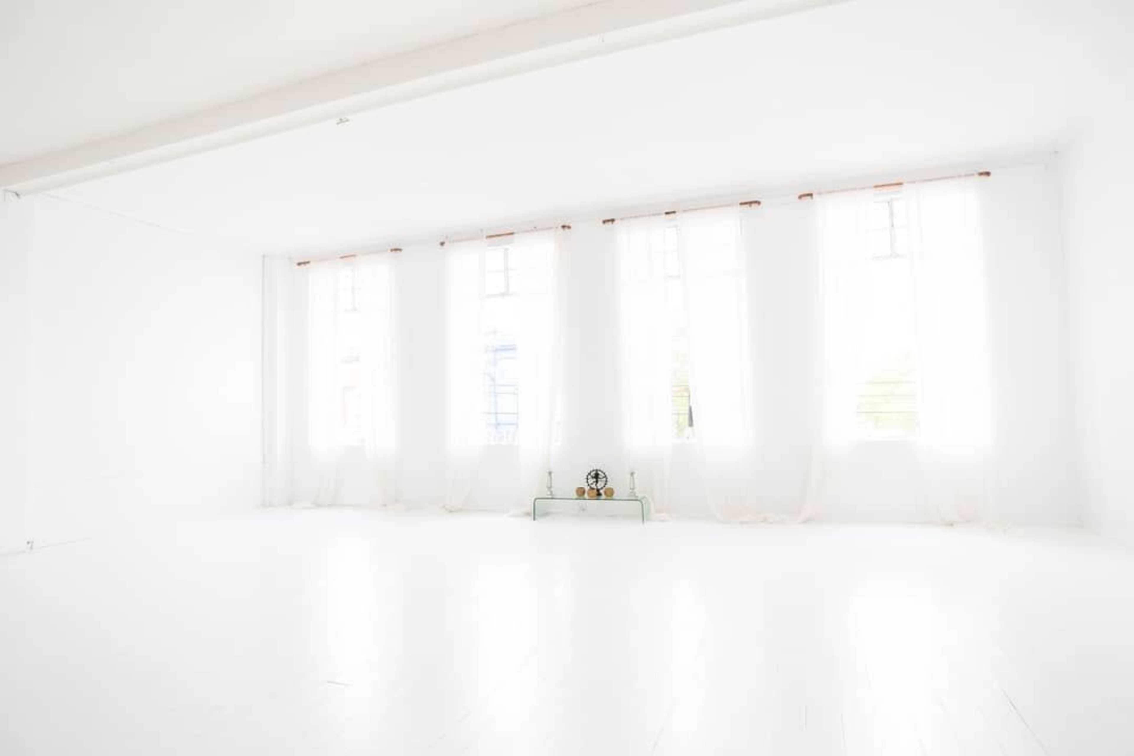 The image shows a bright, empty room with white walls, a white floor, and large windows covered with sheer curtains.