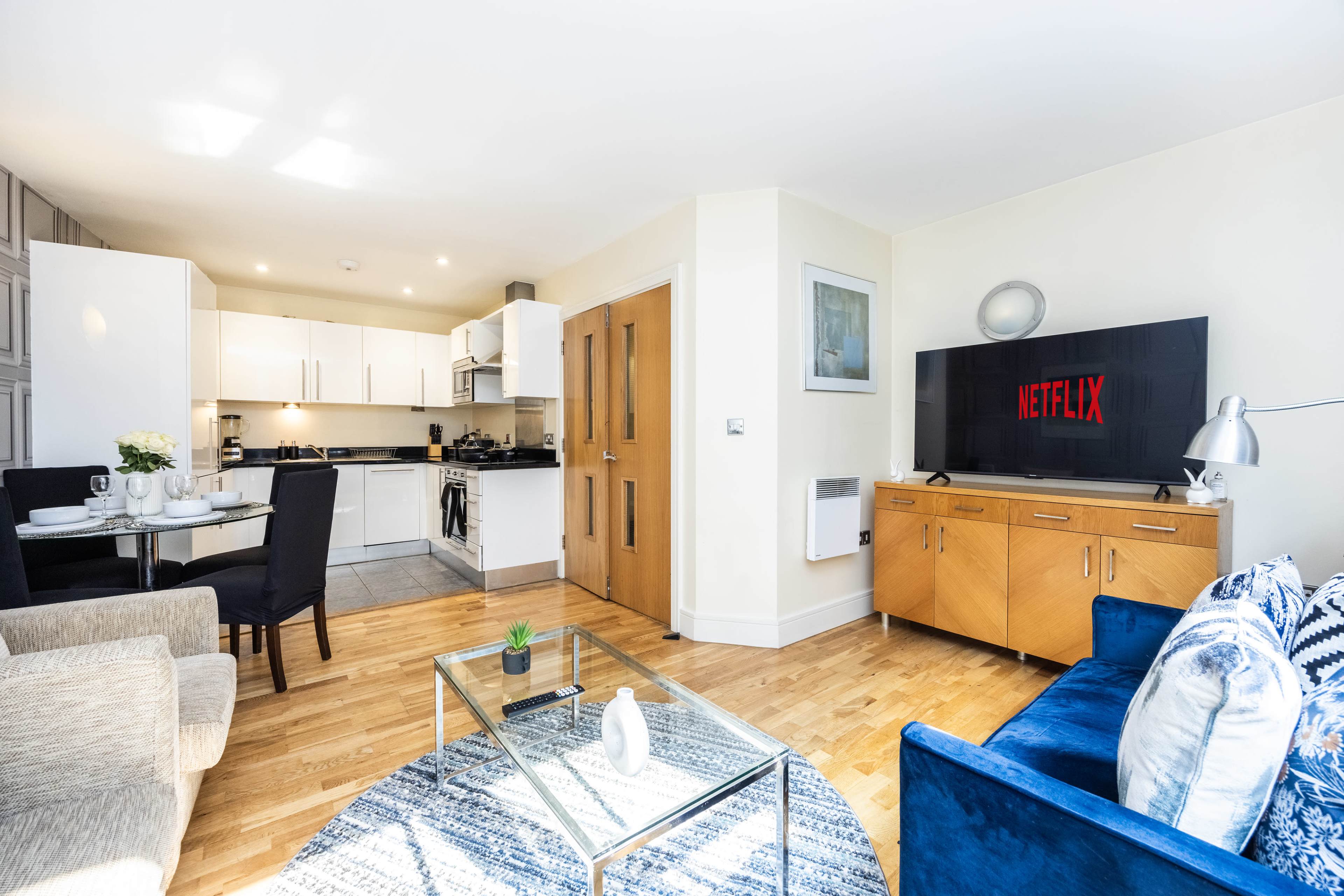 The image shows a modern open-plan living area with a kitchen, dining table, and entertainment unit displaying a "NETFLIX" logo.