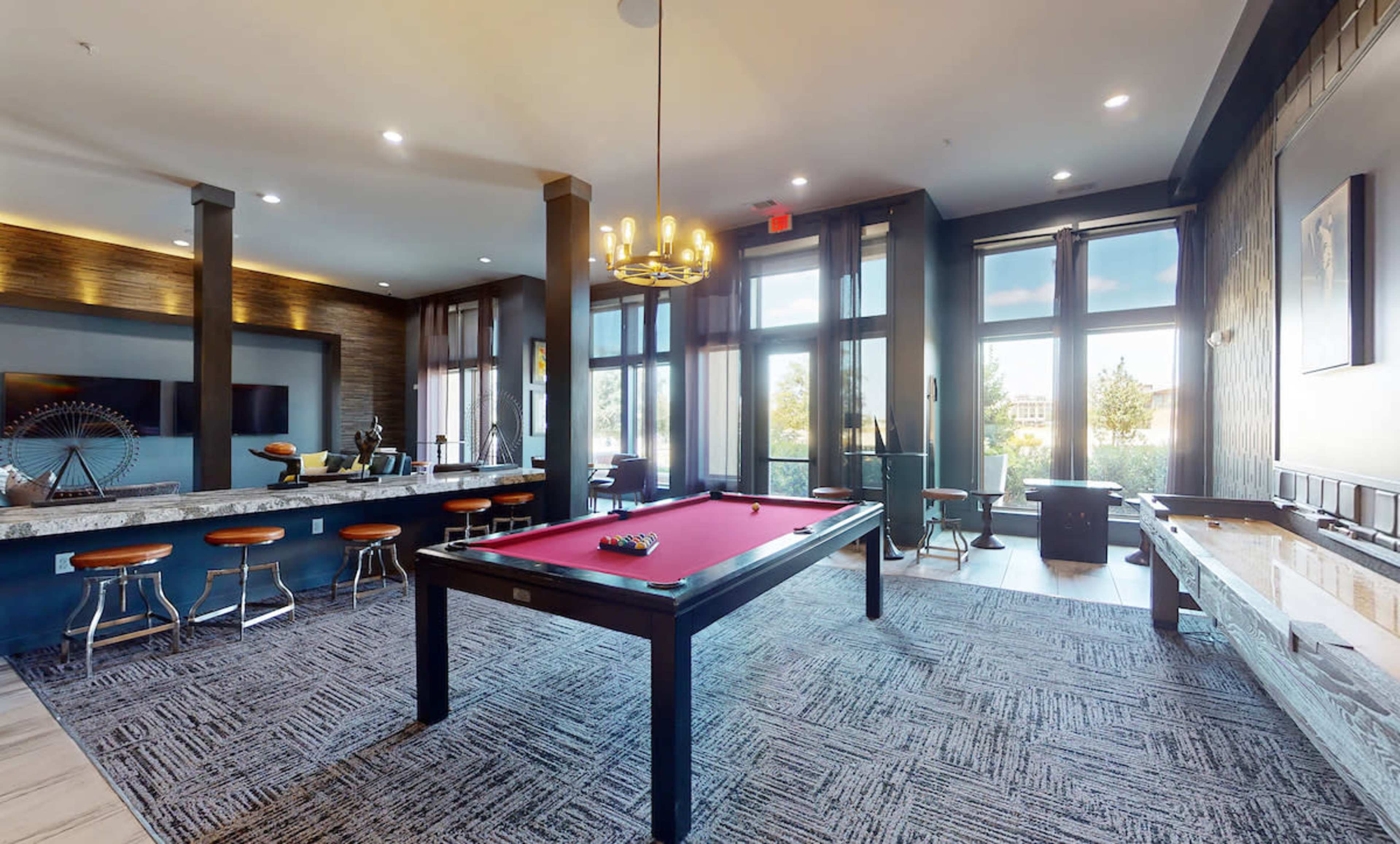 A spacious game room featuring a red pool table, a bar area with stools, and large windows allowing natural light to fill the space.