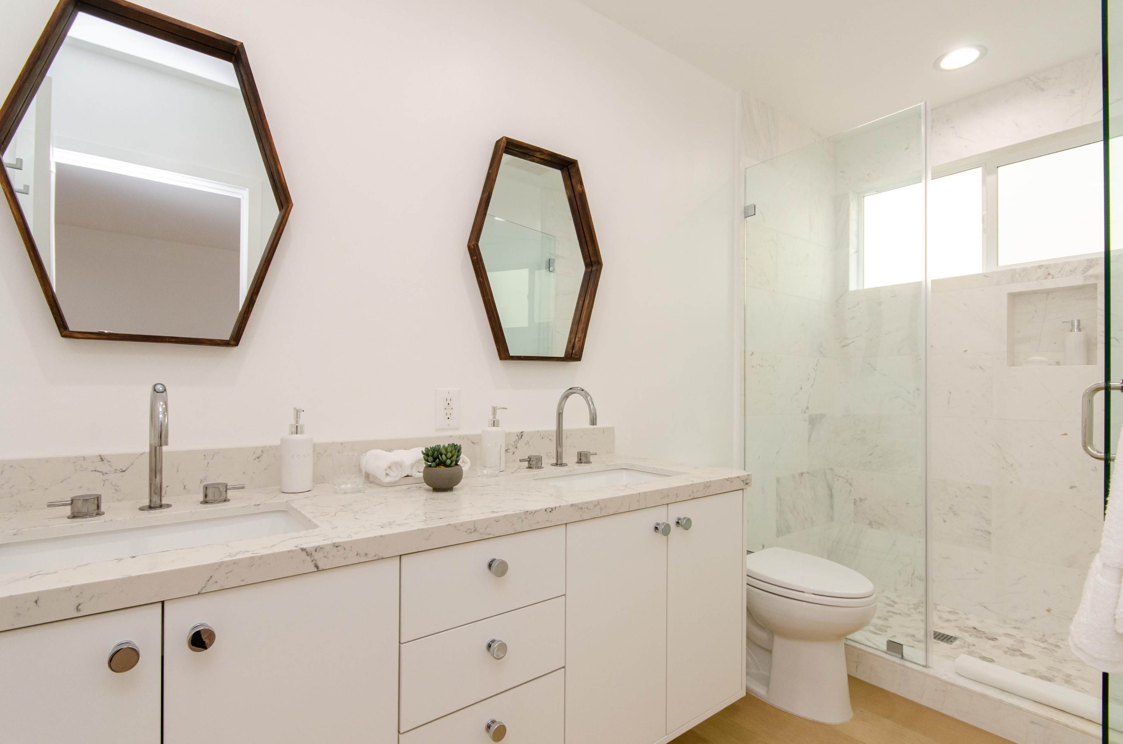 A modern bathroom features a double sink vanity with hexagonal mirrors, a glass shower enclosure, and neutral-toned marble countertops.