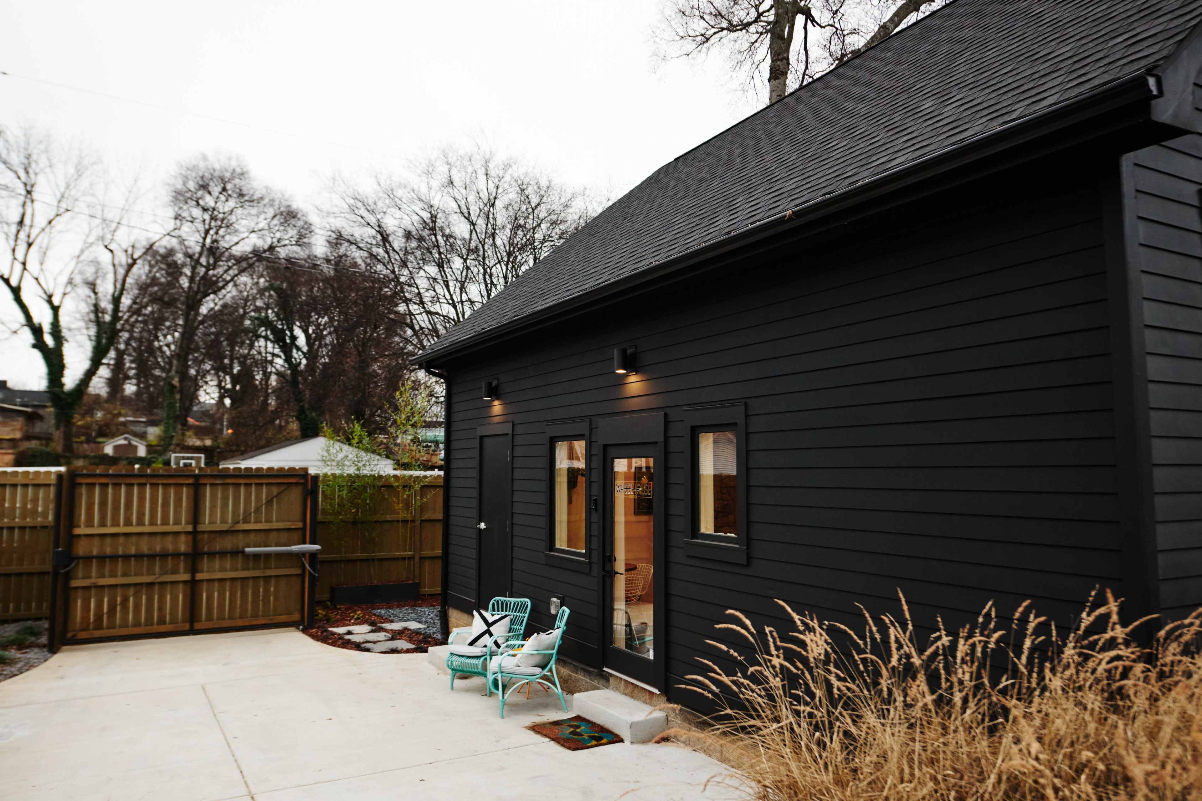 A black wood-sided house features a small patio area with a couple of light blue chairs and a fenced yard in the background.
