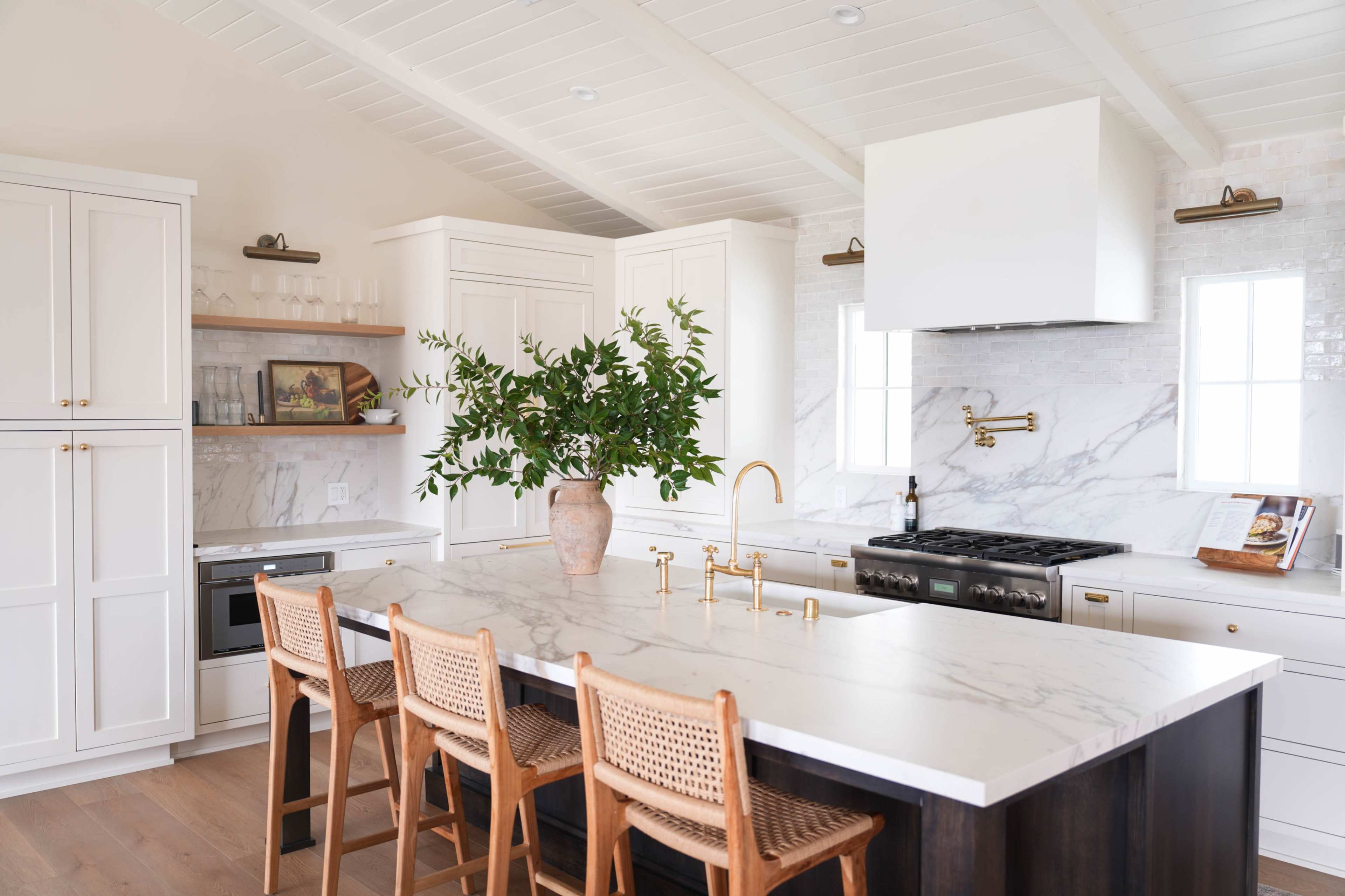 A bright kitchen features a large marble countertop with four wooden stools, white cabinetry, and a stainless steel stove against a marble backsplash.