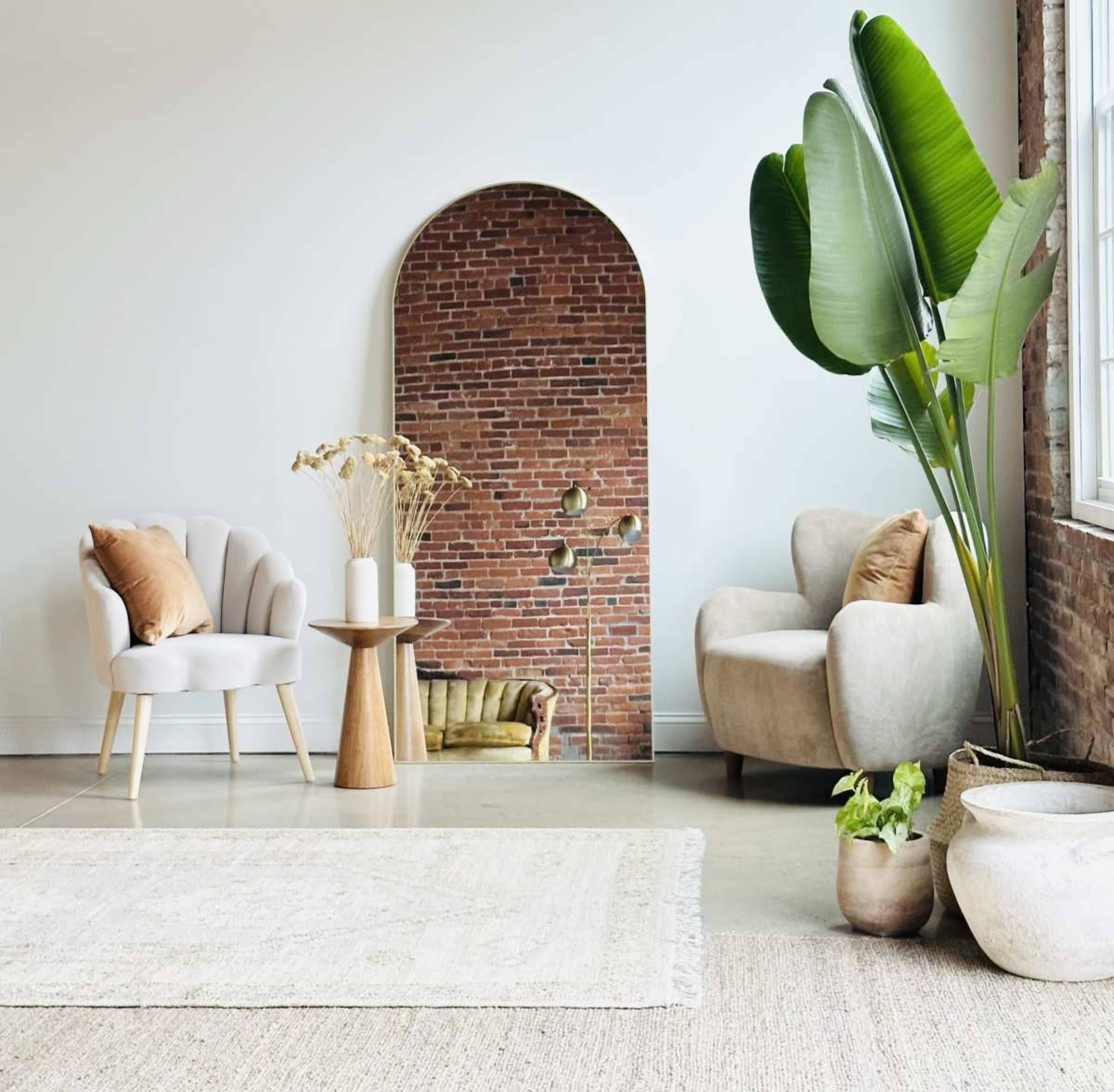 The image shows a modern living space featuring two comfortable chairs, a round wooden table with a vase, a potted plant, and a mirror framed by a brick wall.