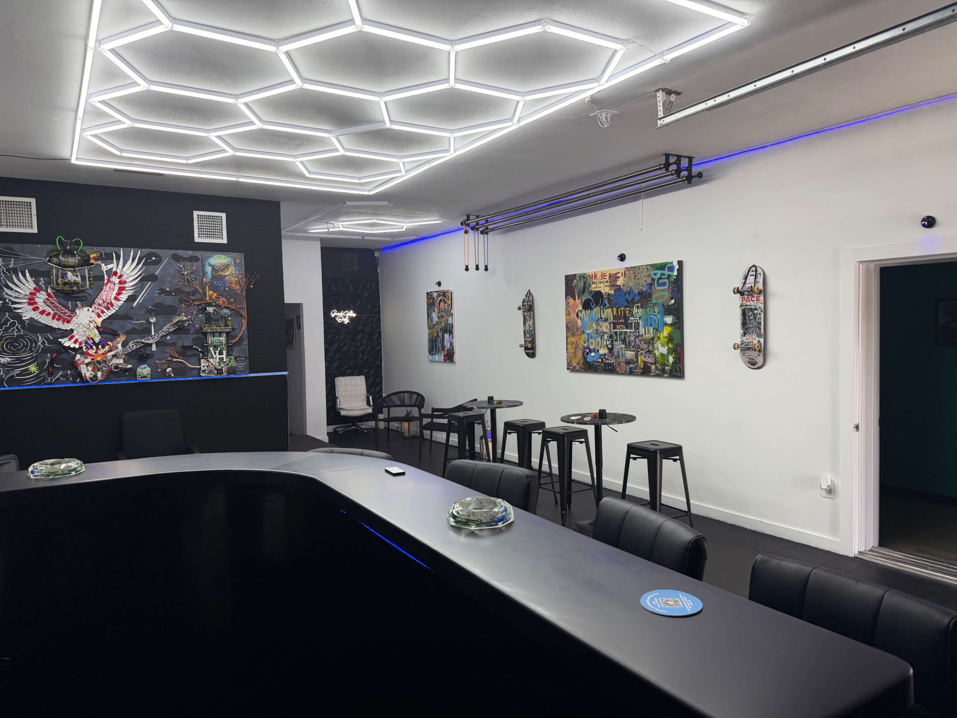 The image shows a modern room with a hexagon-patterned ceiling, a large black counter, wall art, and several high-top tables with chairs.