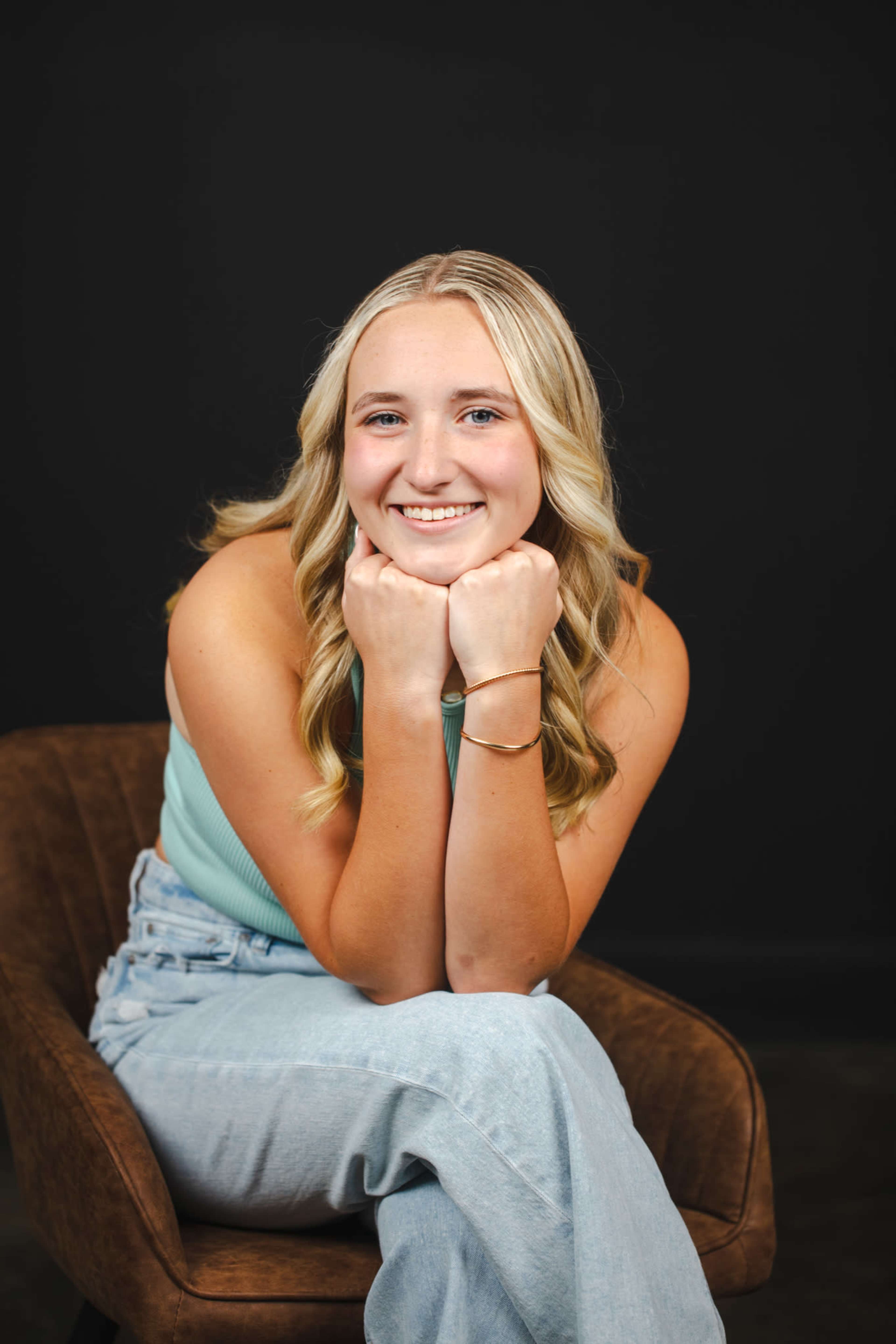 A young woman with blonde hair is sitting in a brown armchair, resting her chin on her hands and smiling.