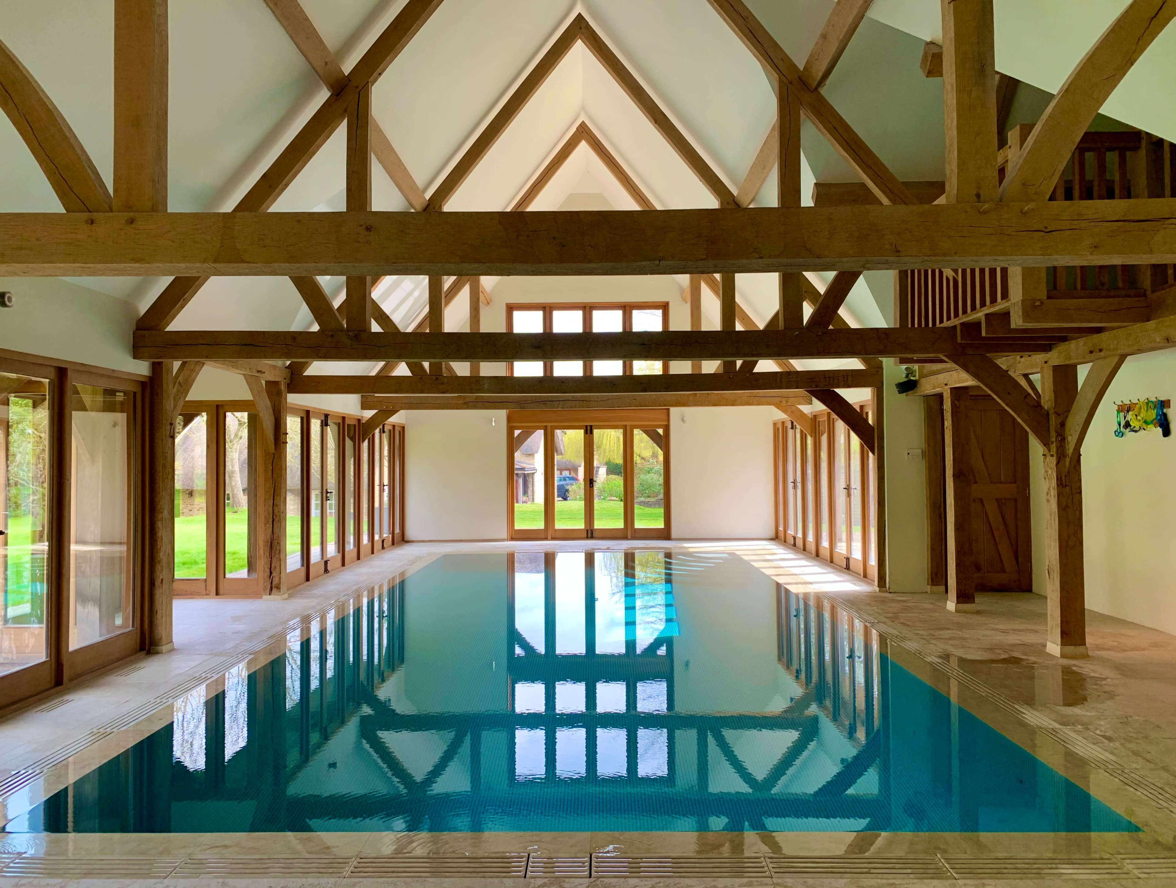 A large indoor swimming pool surrounded by wooden beams and glass doors, with natural light illuminating the space.