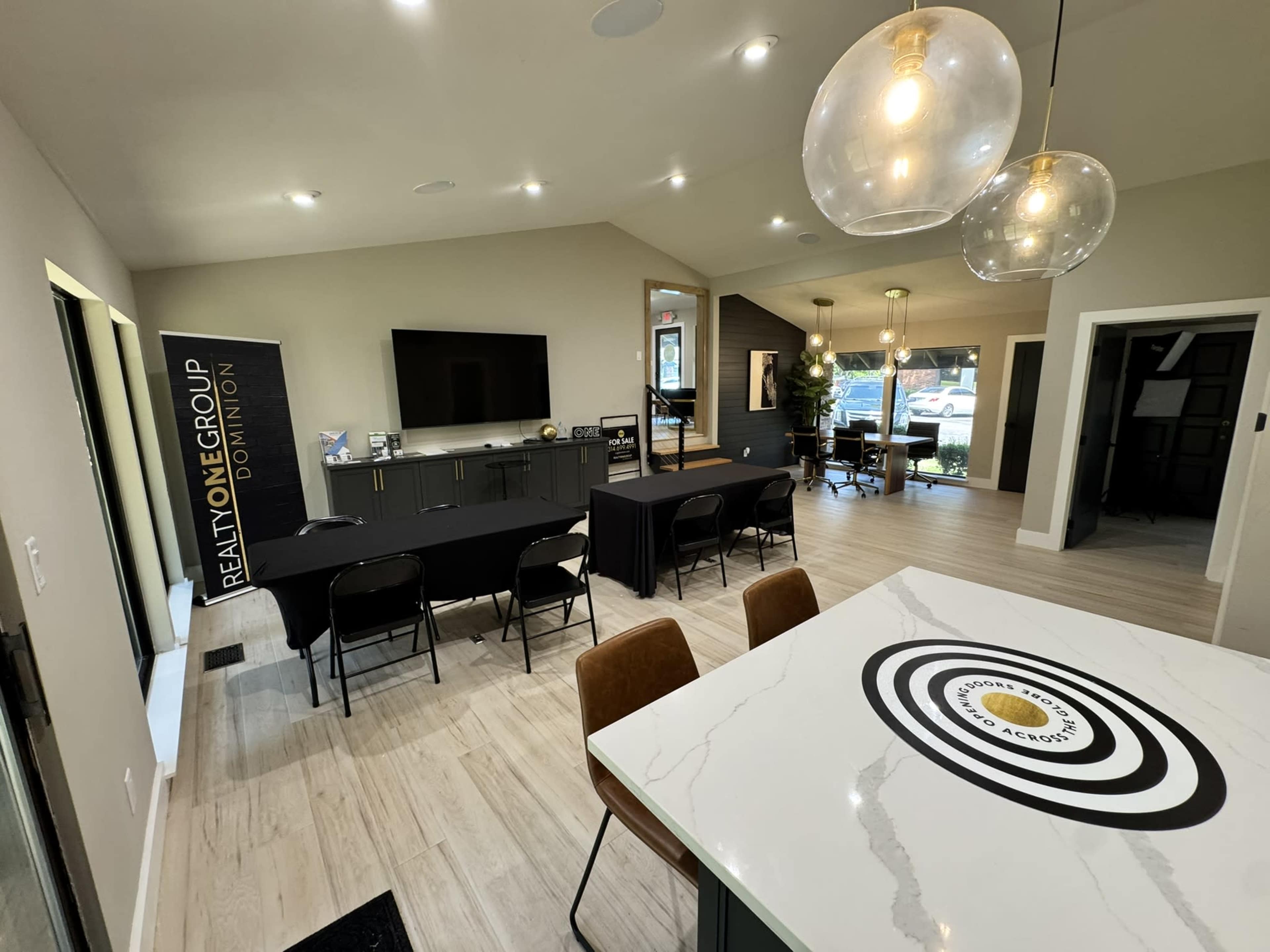 The image shows a modern, open-concept office space with a large television, a reception area, and several tables set up for meetings or presentations.