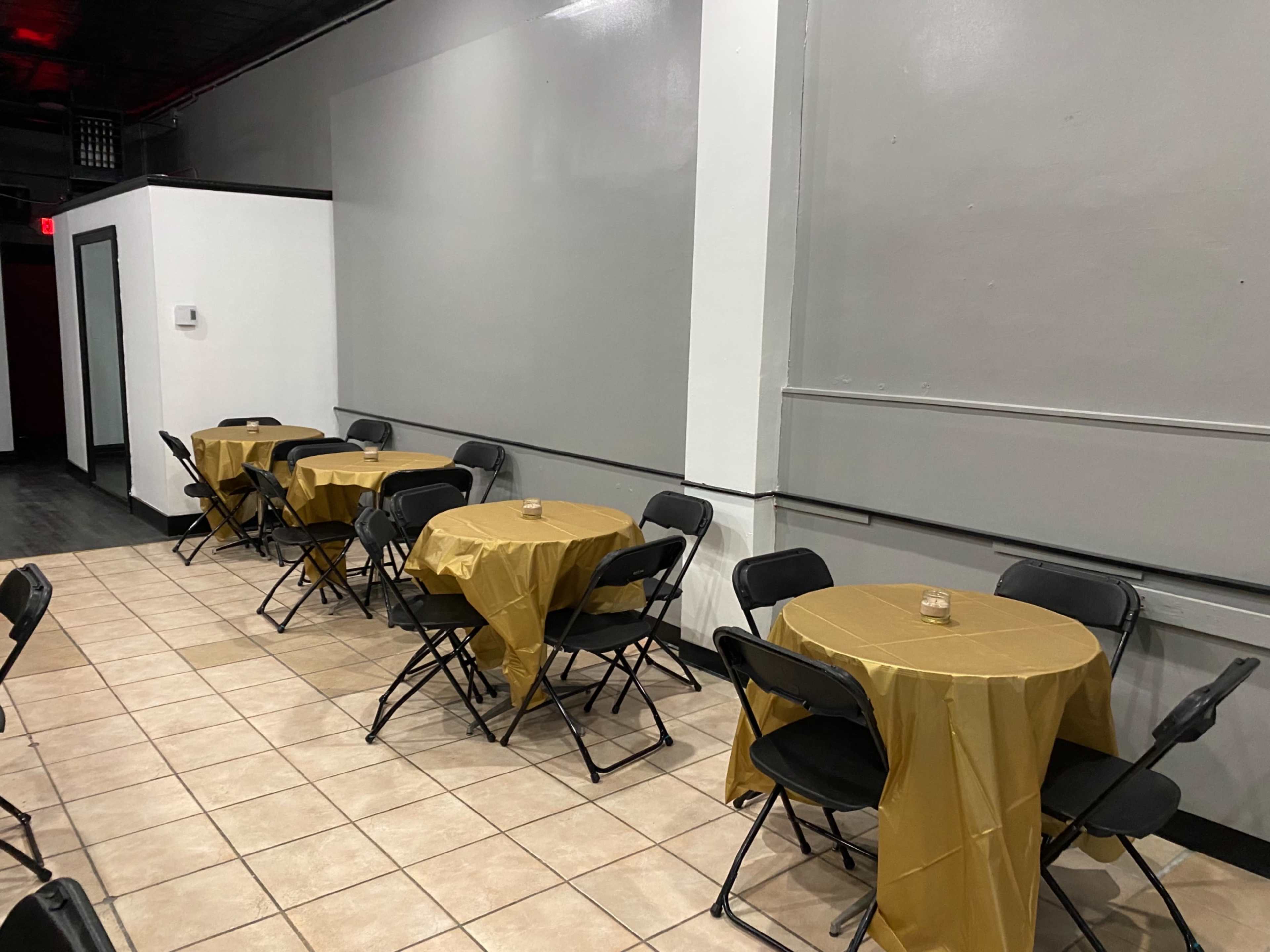 A small indoor event space with several round tables covered in gold tablecloths and surrounded by black folding chairs.