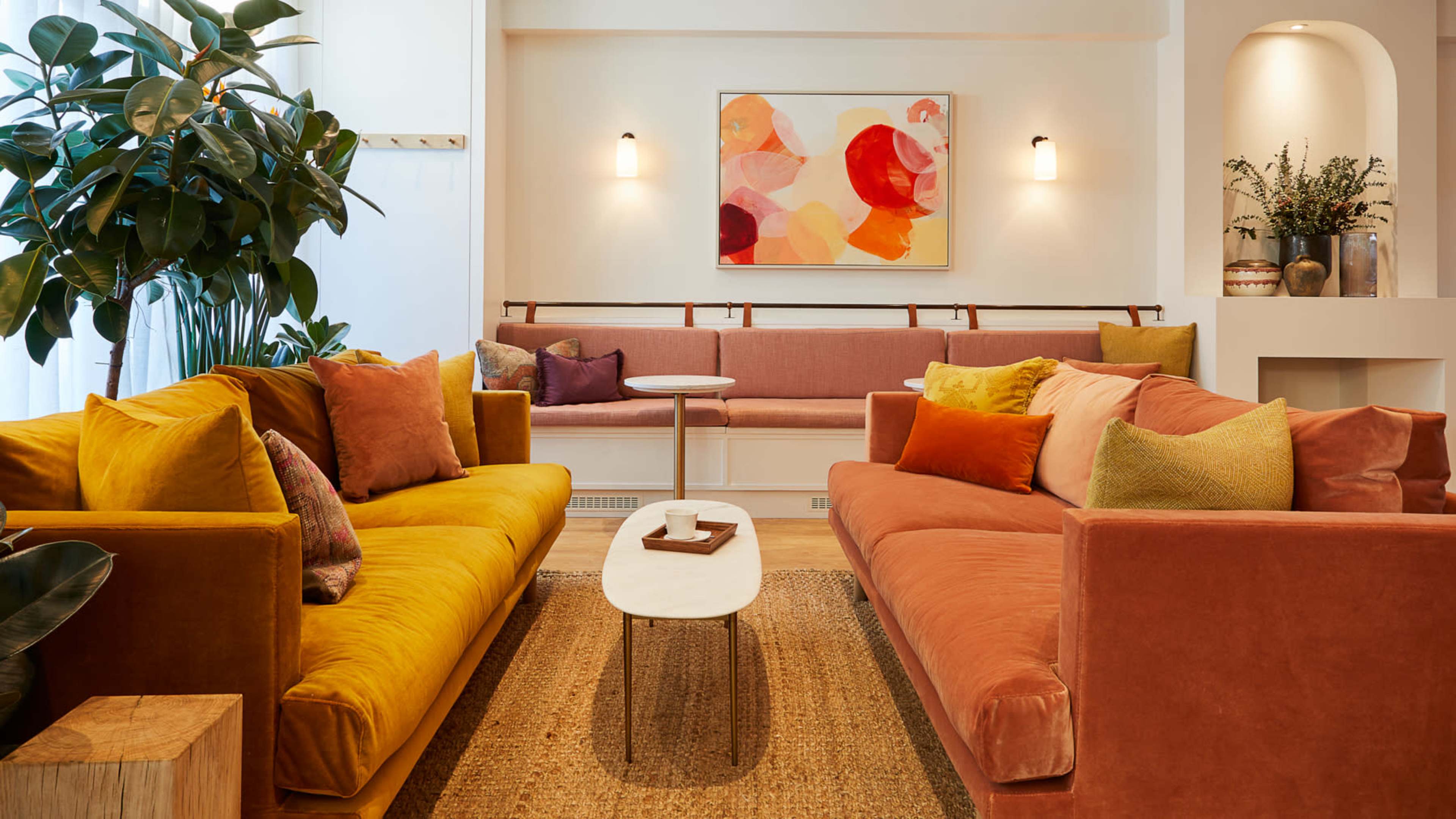 The image shows a cozy lounge area featuring two upholstered sofas in warm colors, a small coffee table, and decorative plants, complemented by abstract artwork on the wall.