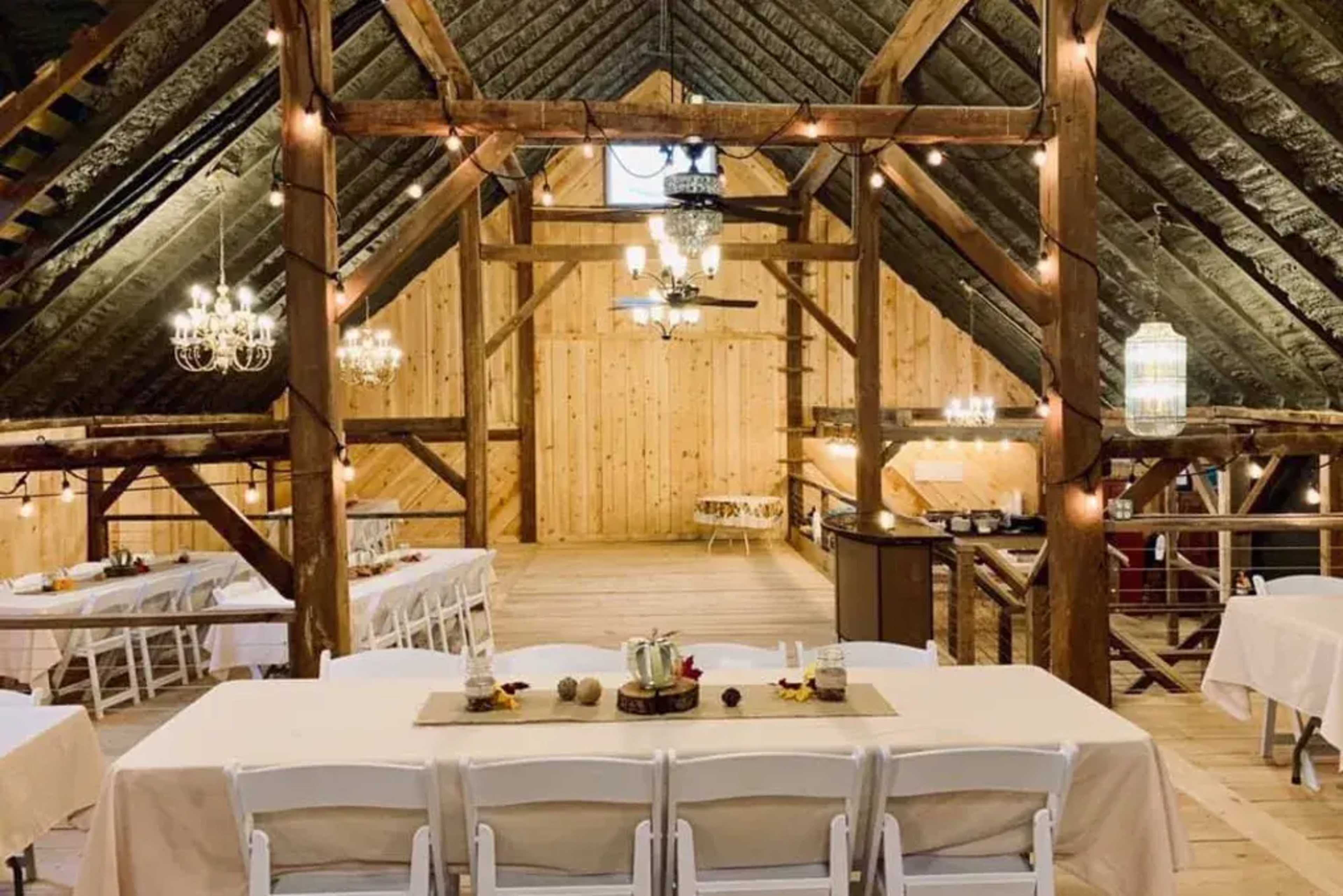 An elegant barn interior with wooden beams, chandeliers, and neatly arranged tables set for an event.