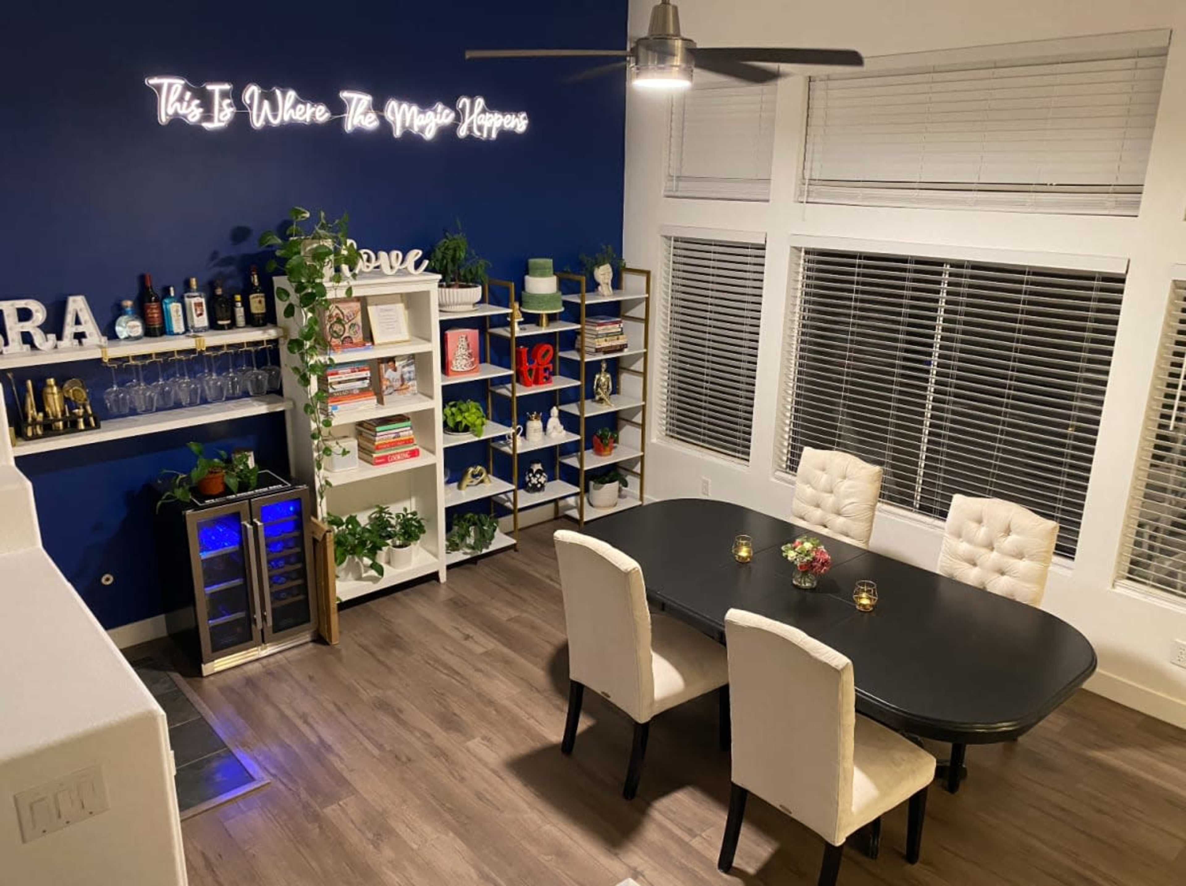 The image shows a modern dining area featuring a black table with white chairs, illuminated by wall decor that reads "This Is Where The Magic Happens," along with shelves displaying books and plants against a dark blue wall.