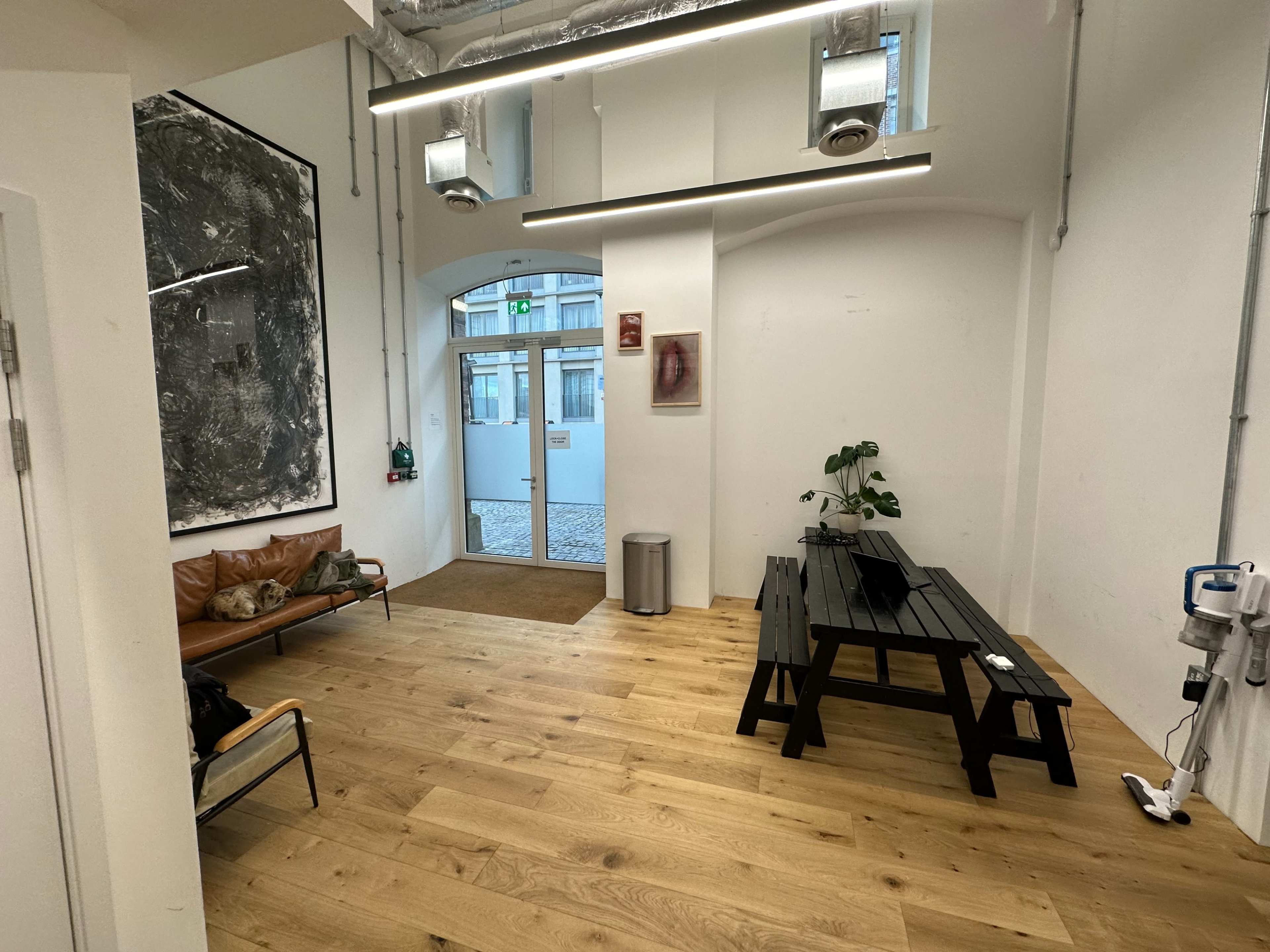 A modern interior space featuring a leather sofa, a wooden table, a potted plant, and large windows with light streaming in.