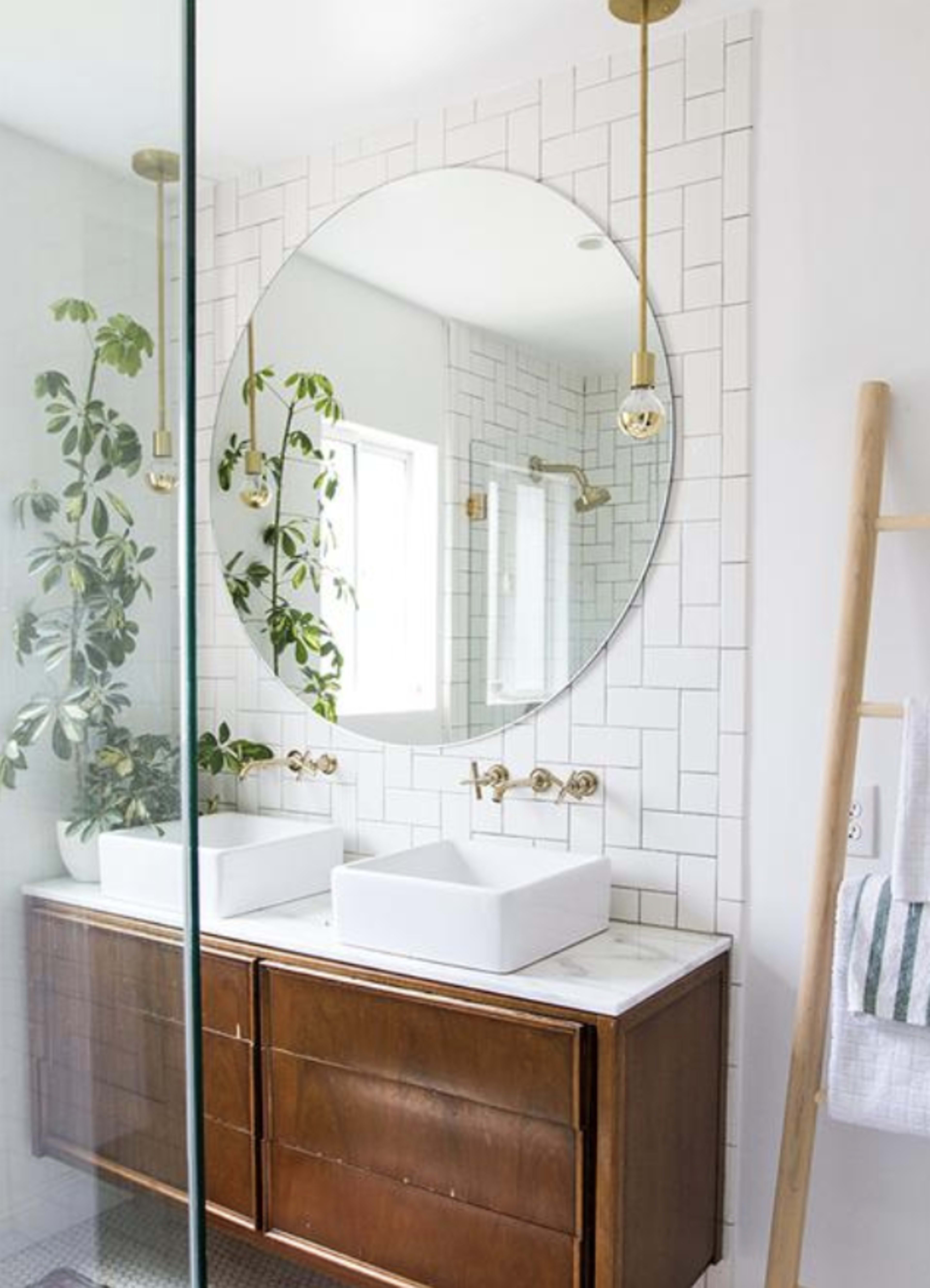 A modern bathroom features a double sink with two white vessel basins, a circular mirror, and a wooden vanity, surrounded by tiled walls and indoor plants.