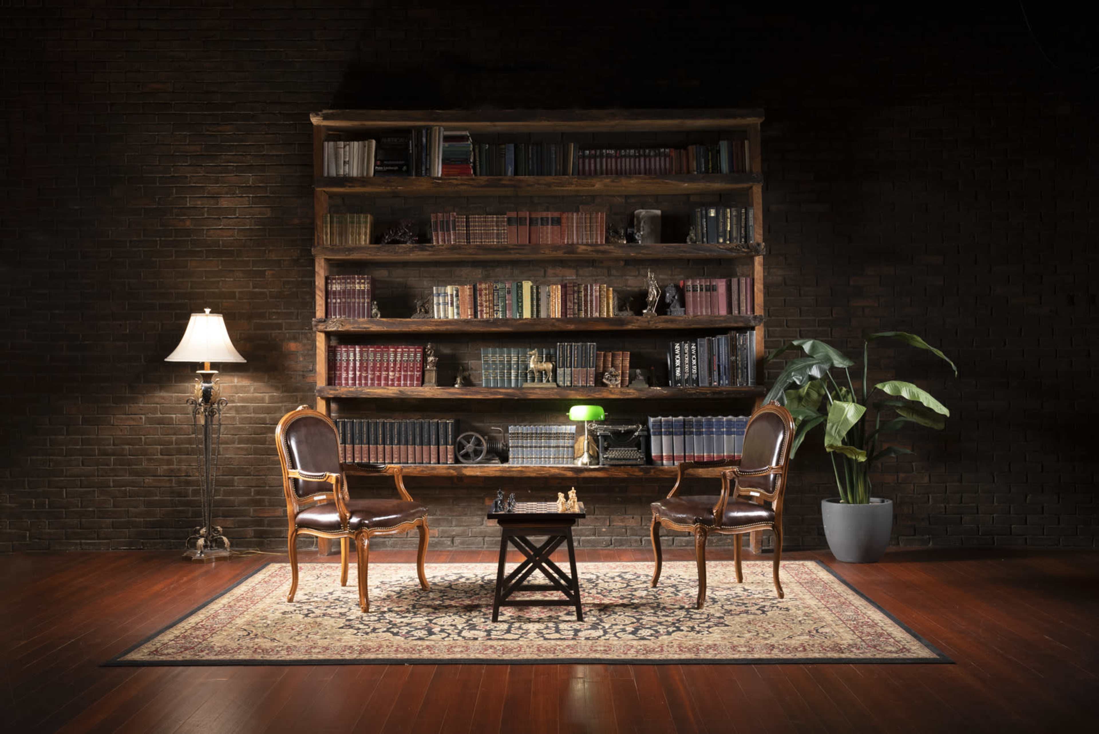 A cozy reading nook features two chairs facing a small table, illuminated by a lamp, with a bookshelf filled with books and a decorative plant nearby.