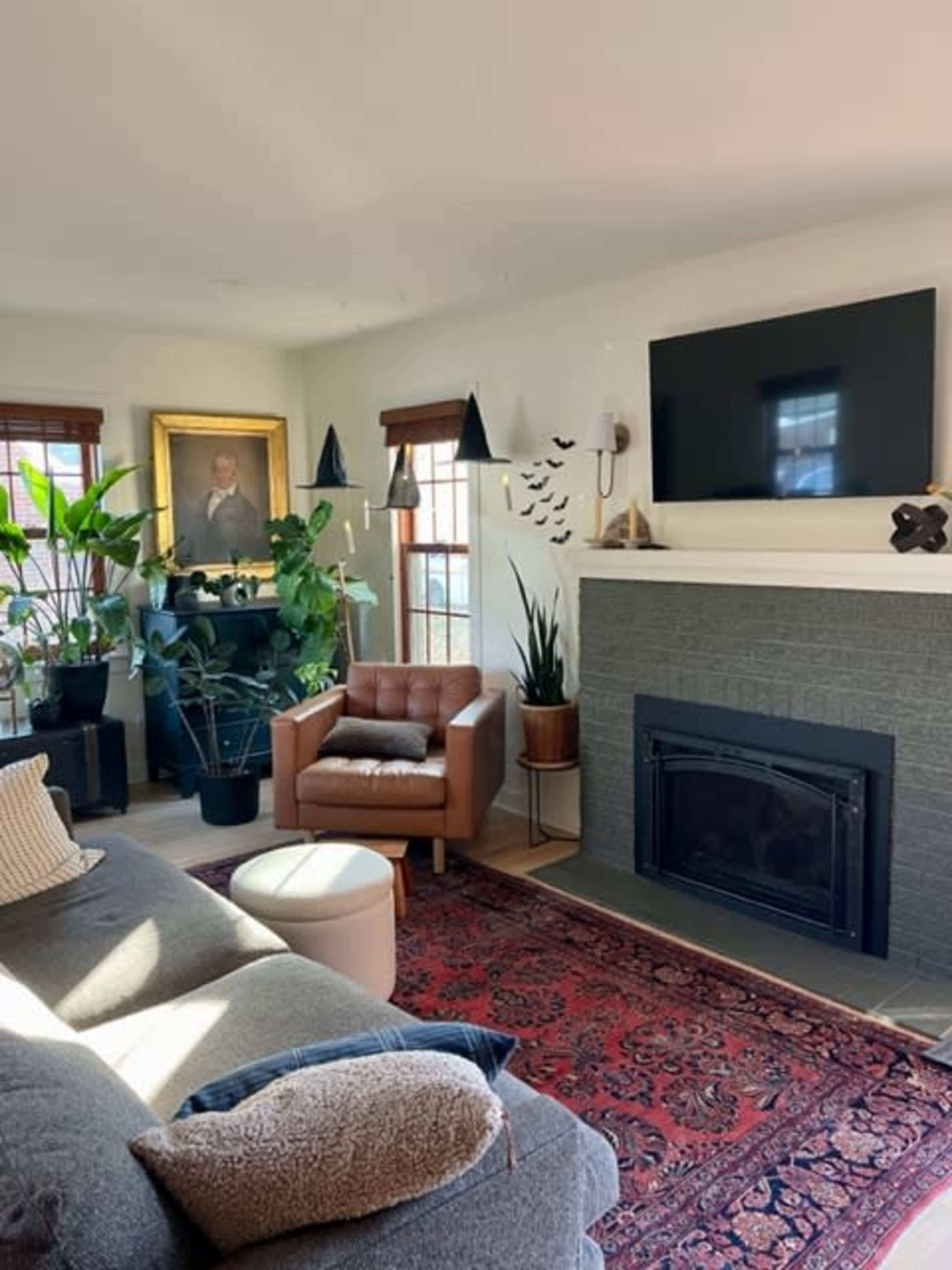 A cozy living room features a brown leather armchair, a large painting, various indoor plants, a contemporary fireplace, and a wall-mounted television.