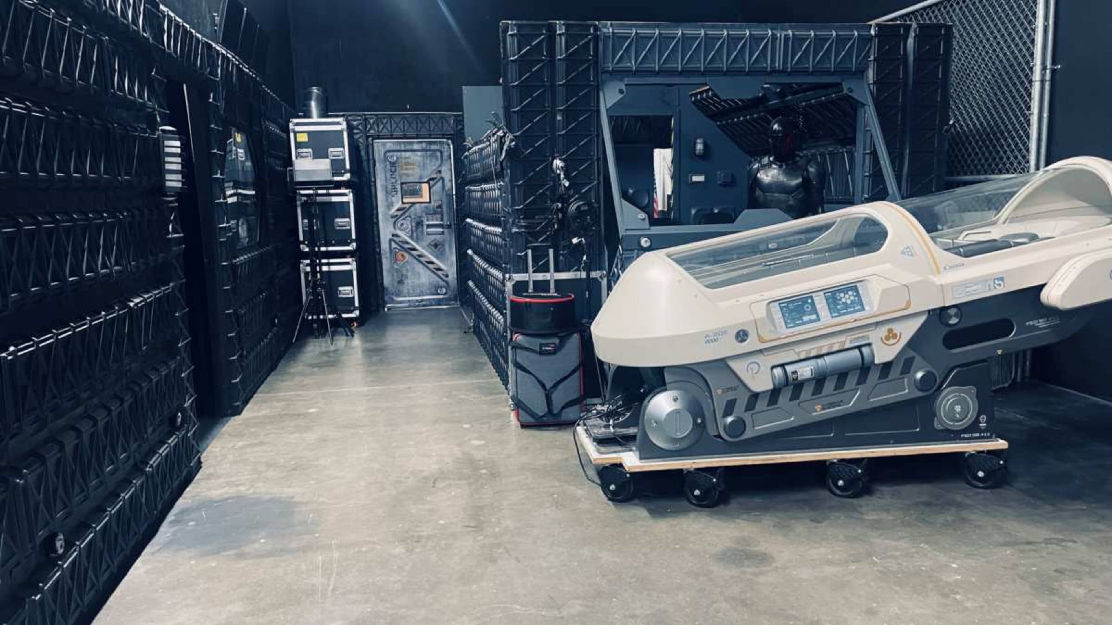 The image shows a dimly lit room containing various industrial-looking equipment and large storage containers along the walls.
