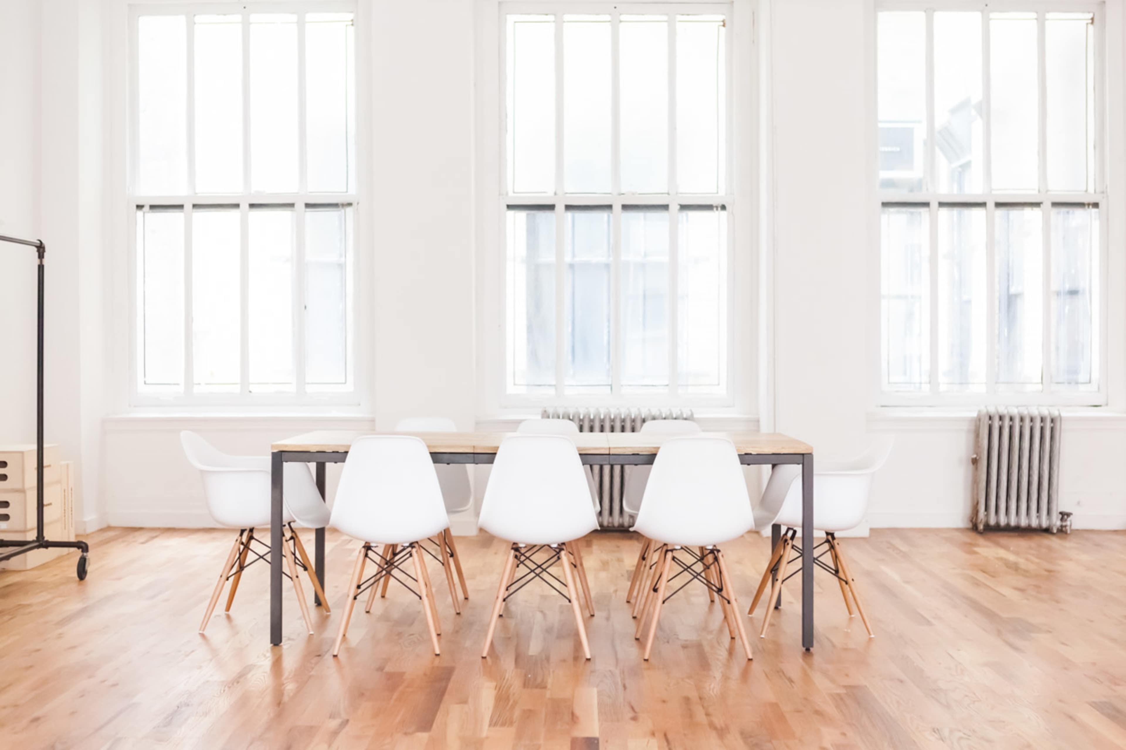 A long wooden table with six white chairs is situated in a bright room featuring large windows and wooden flooring.