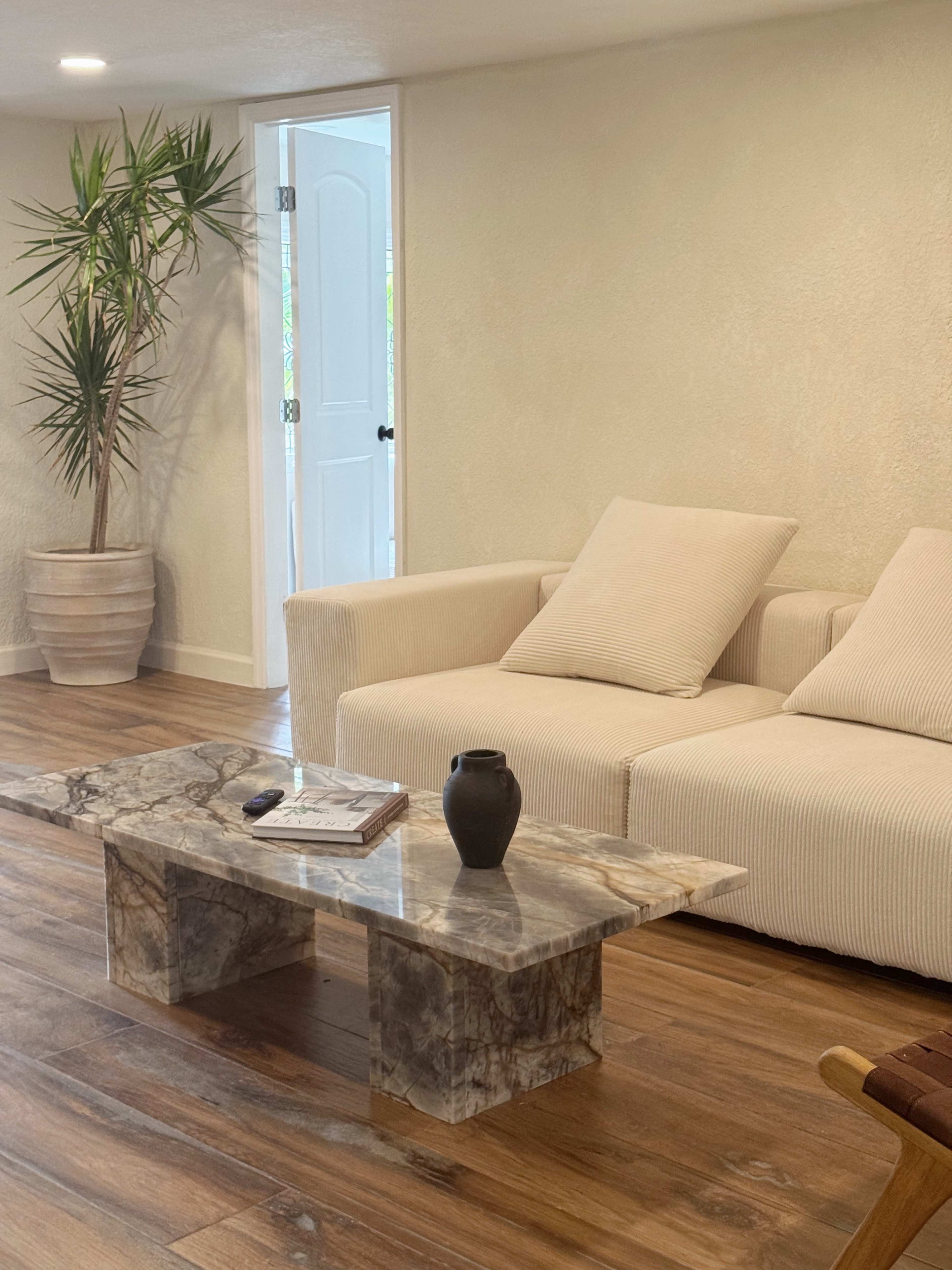 A modern living room features a white sofa, a marble coffee table, and a doorway displaying a potted plant.