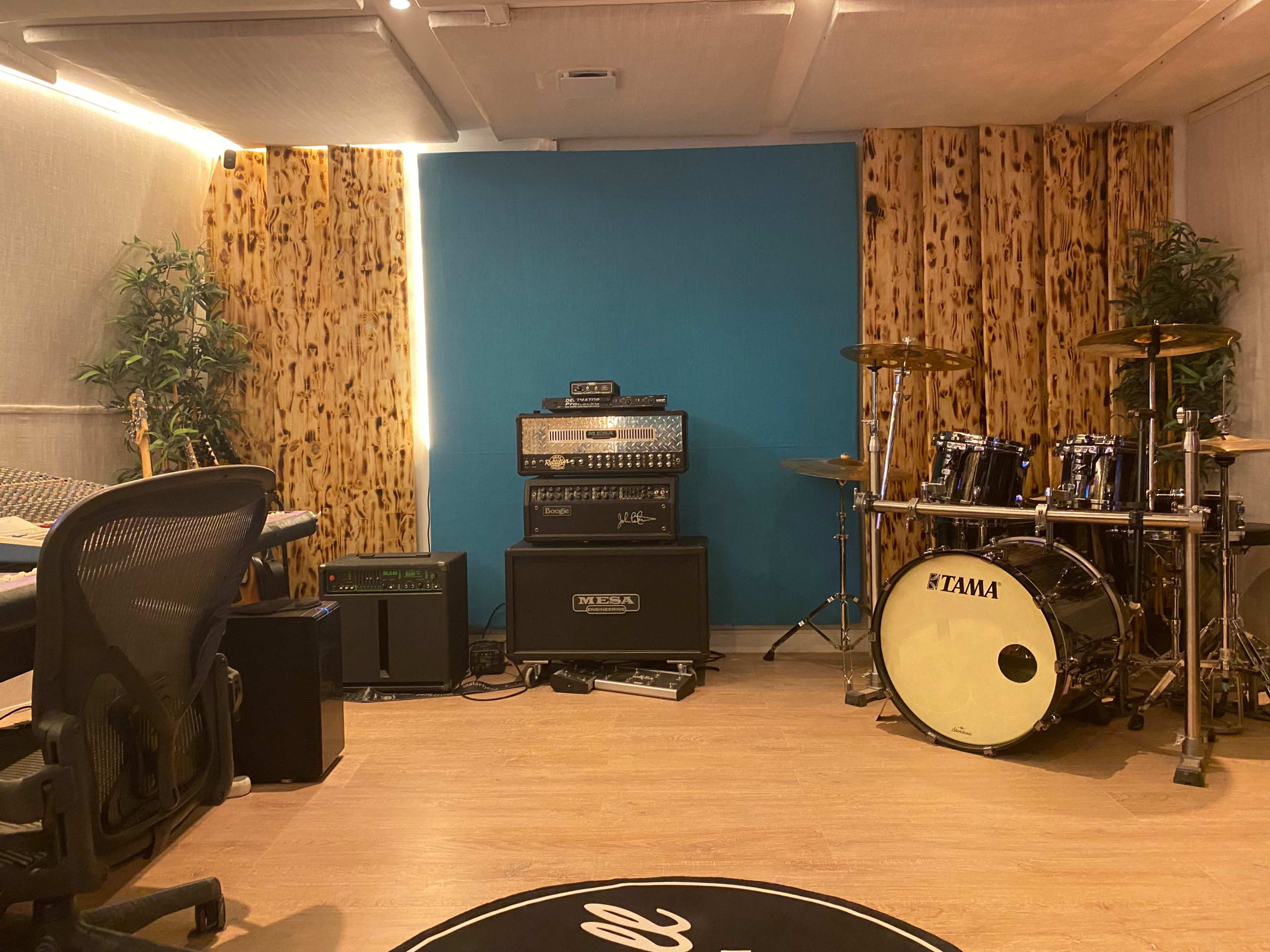 A recording studio with musical instruments, including a drum set, guitar amplifiers, and a desk chair, set against textured wooden walls.