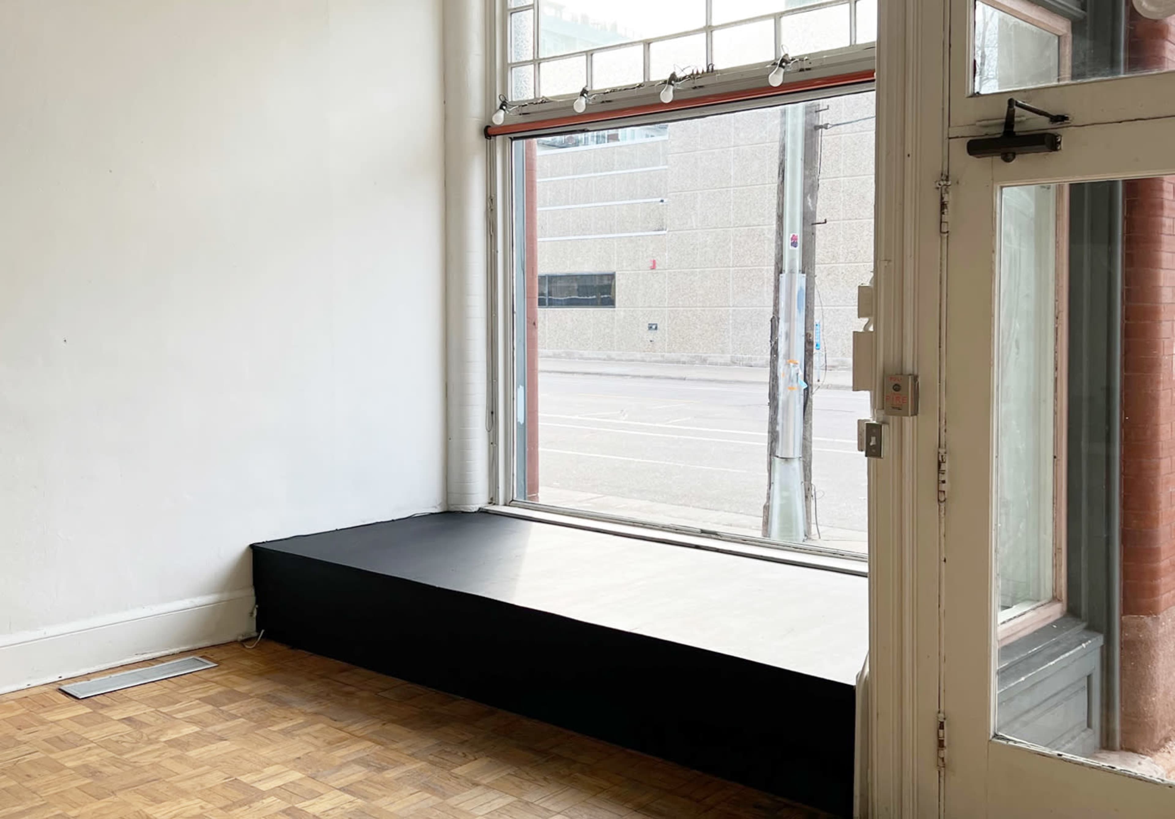 The image shows a vacant corner of a room with a black bench under a large window, revealing a view of a street outside.