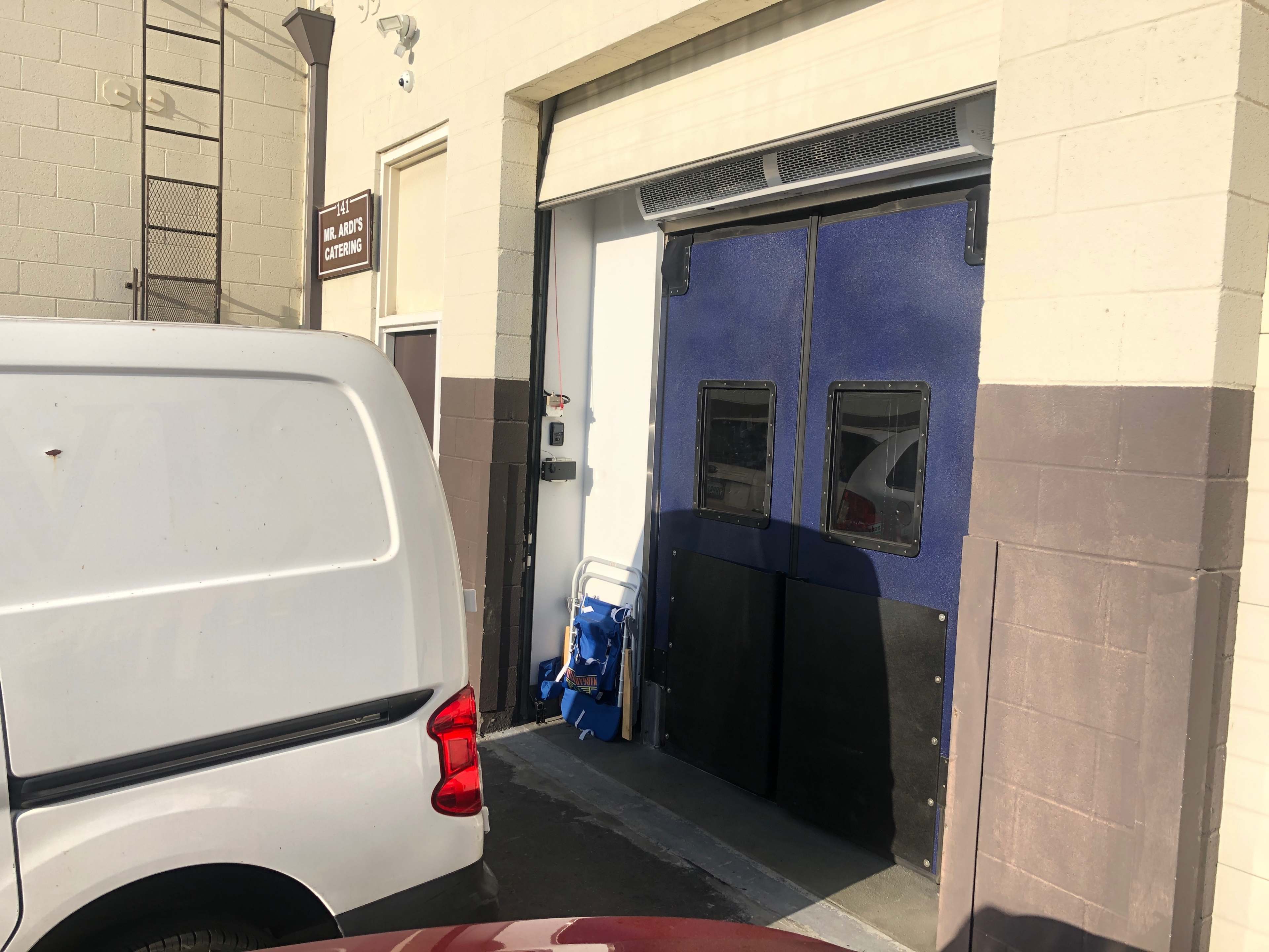 A white van is parked near a blue double door entrance of a building, with a hand truck visible beside the door.