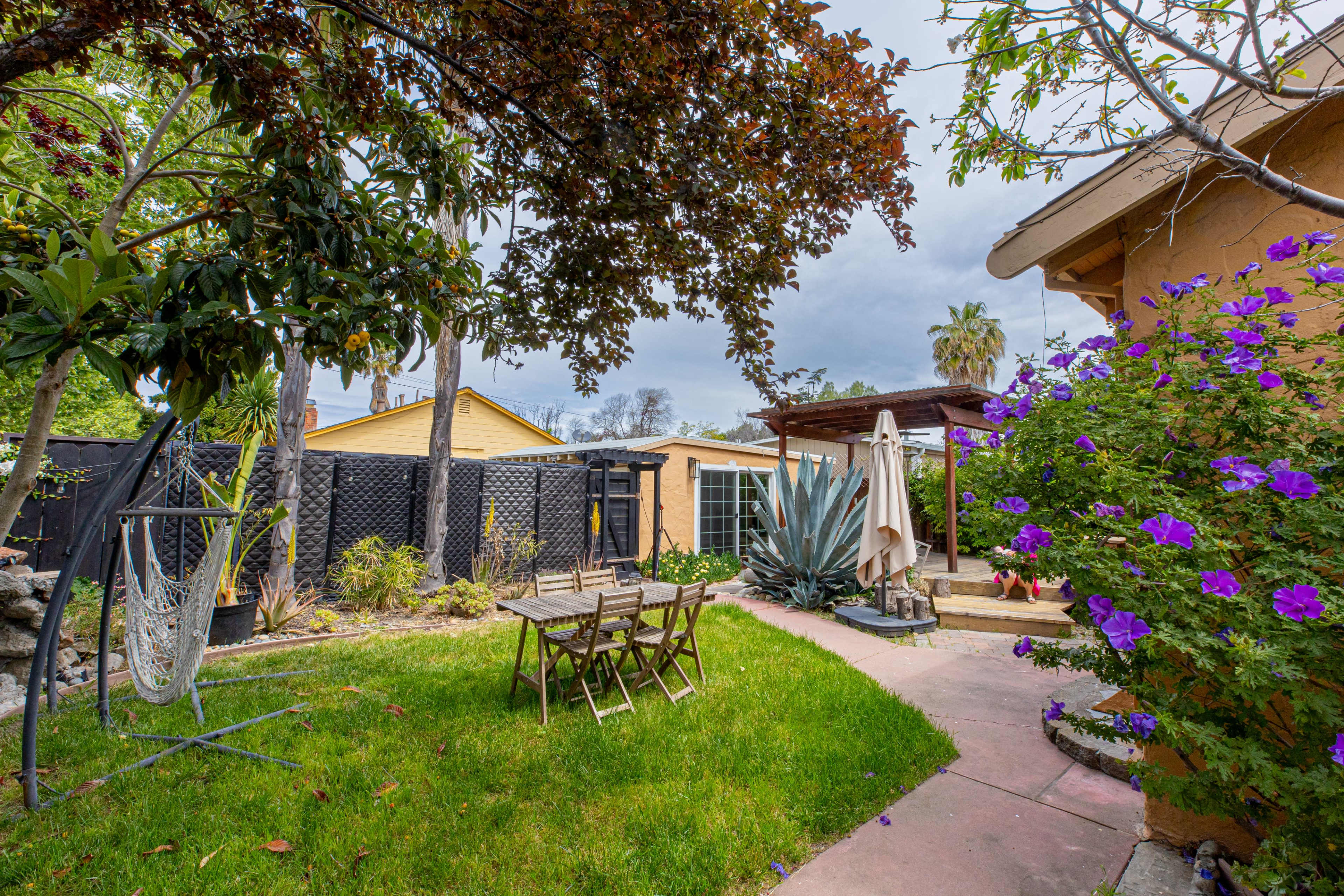 The image features a well-maintained backyard with a grassy area, seating around a small table, and vibrant purple flowers along the pathway.