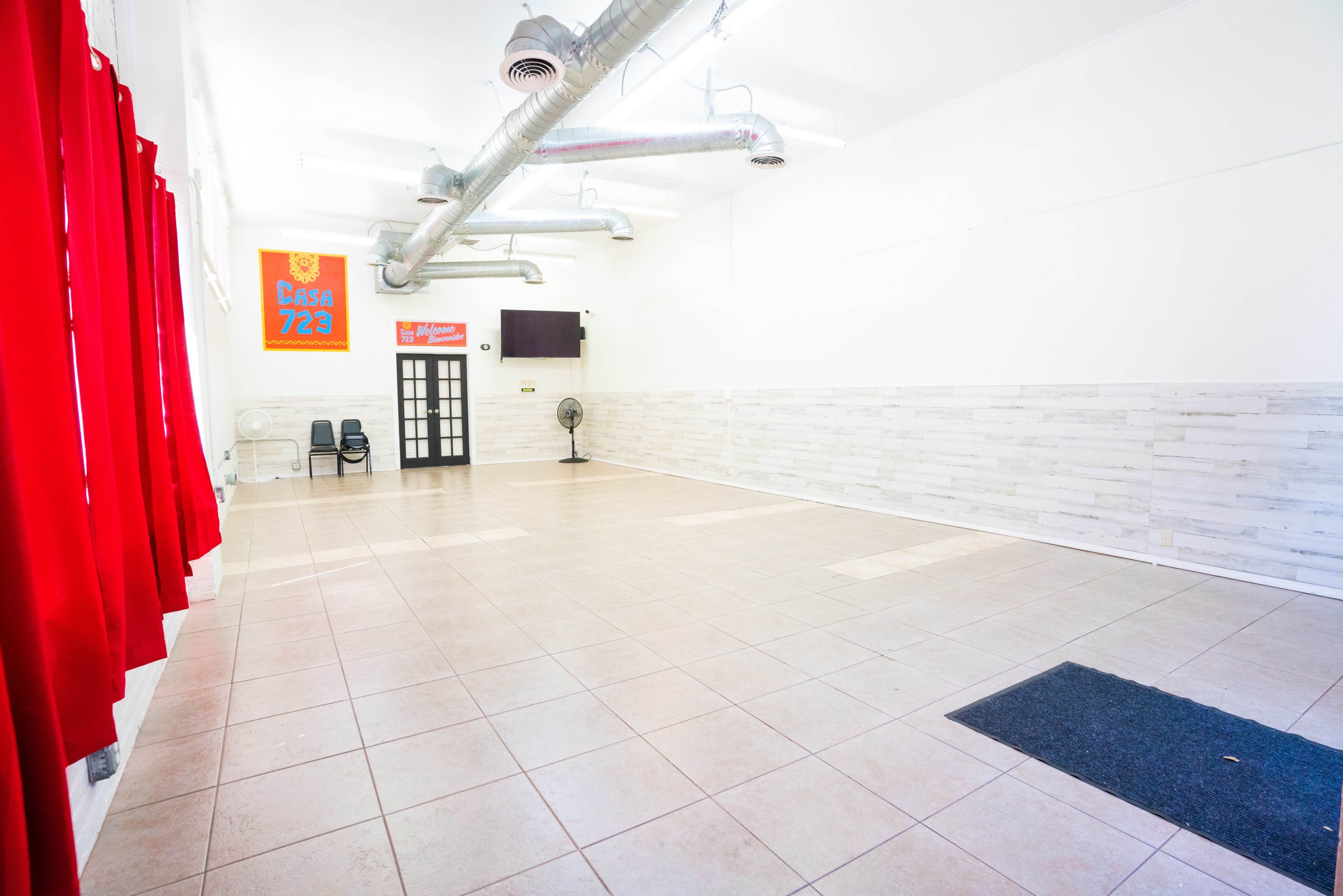 A spacious, empty room with tiled flooring, red curtains on one side, and a door leading to another area, along with a small fan in the corner.