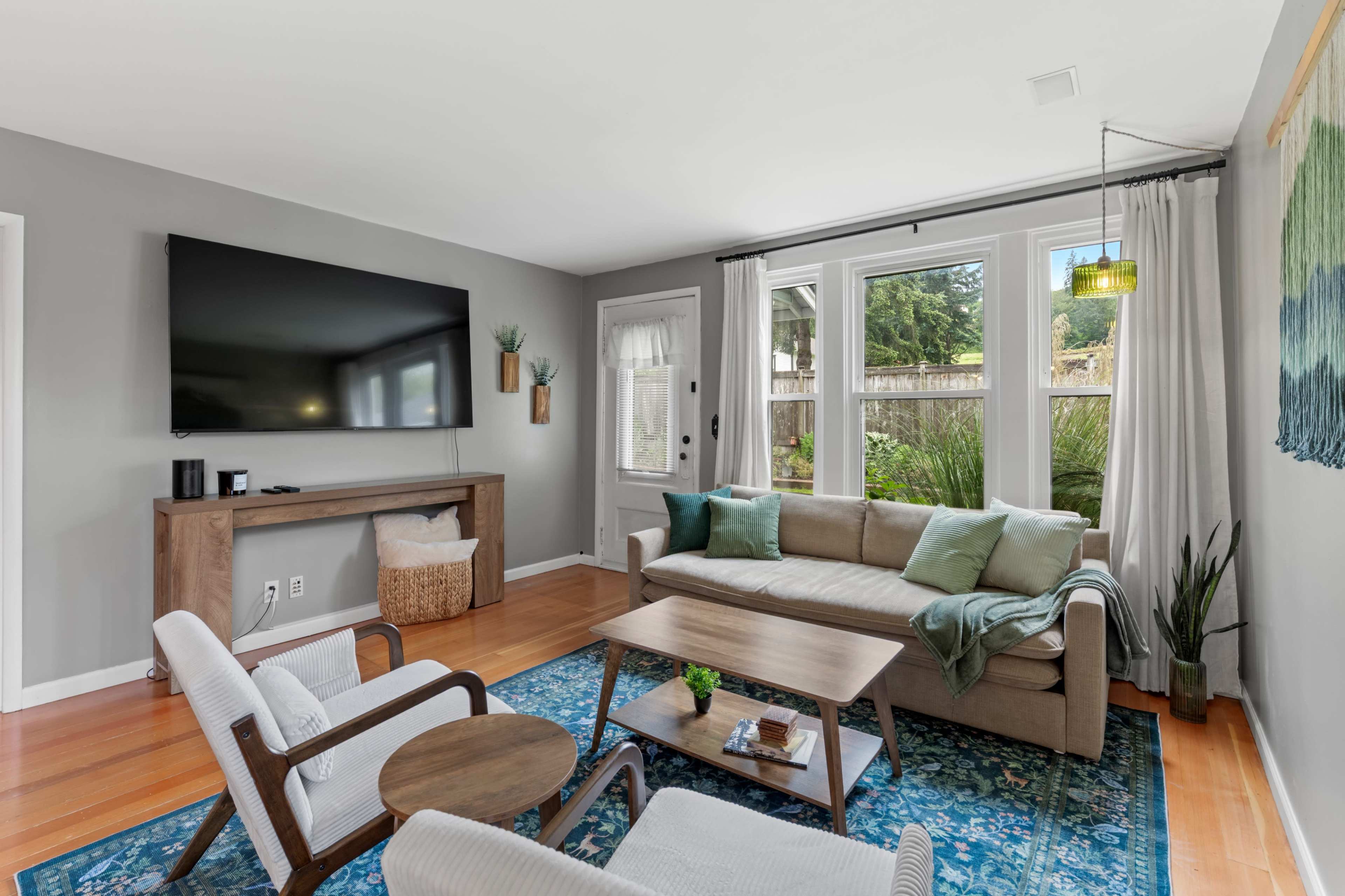 The living room features a comfortable seating arrangement with a large television mounted on the wall, a wooden console table, and bright greenery visible through the windows.
