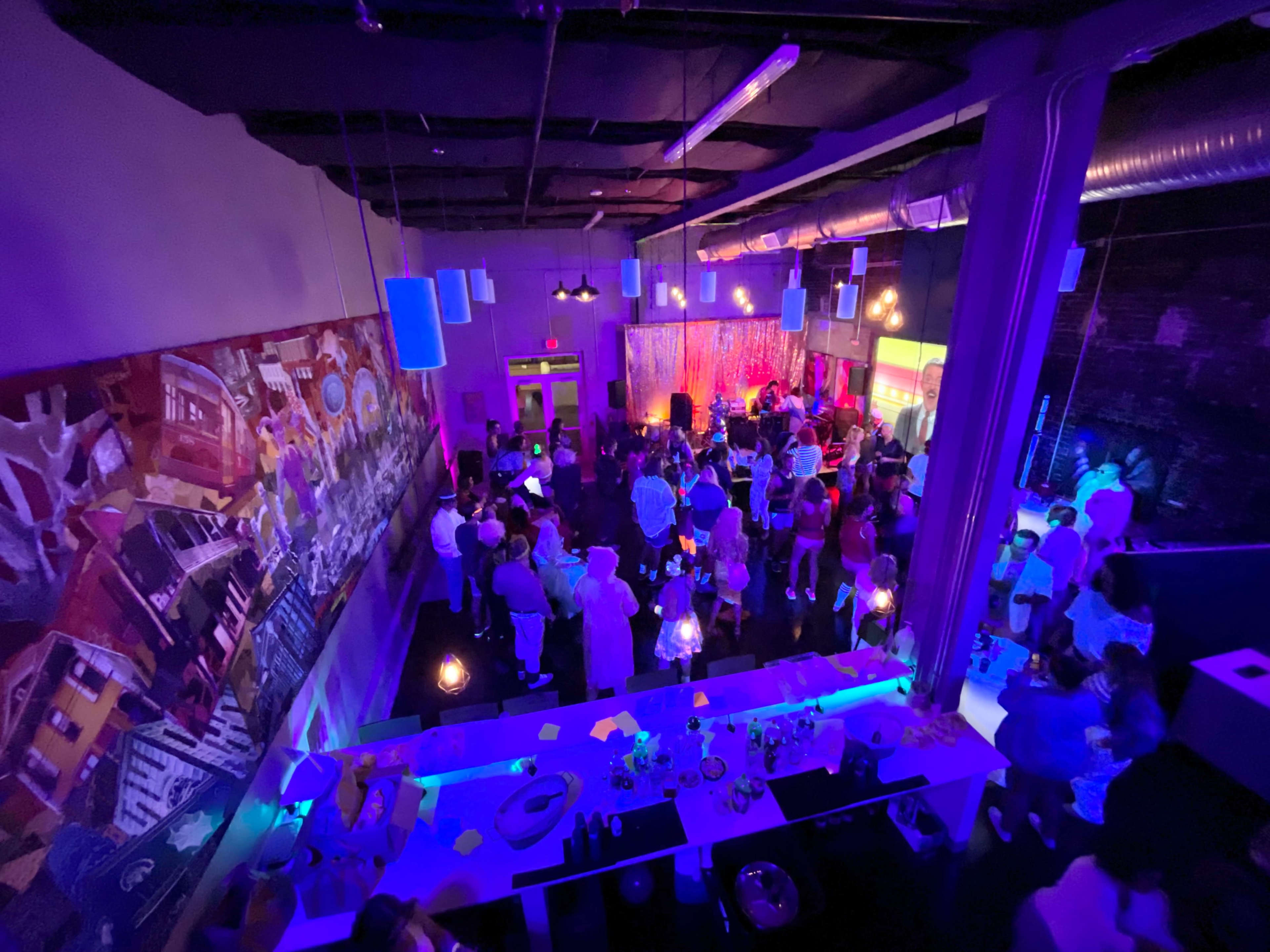 A crowded indoor venue features vibrant lighting and a large mural on the wall, with people socializing and enjoying the atmosphere.