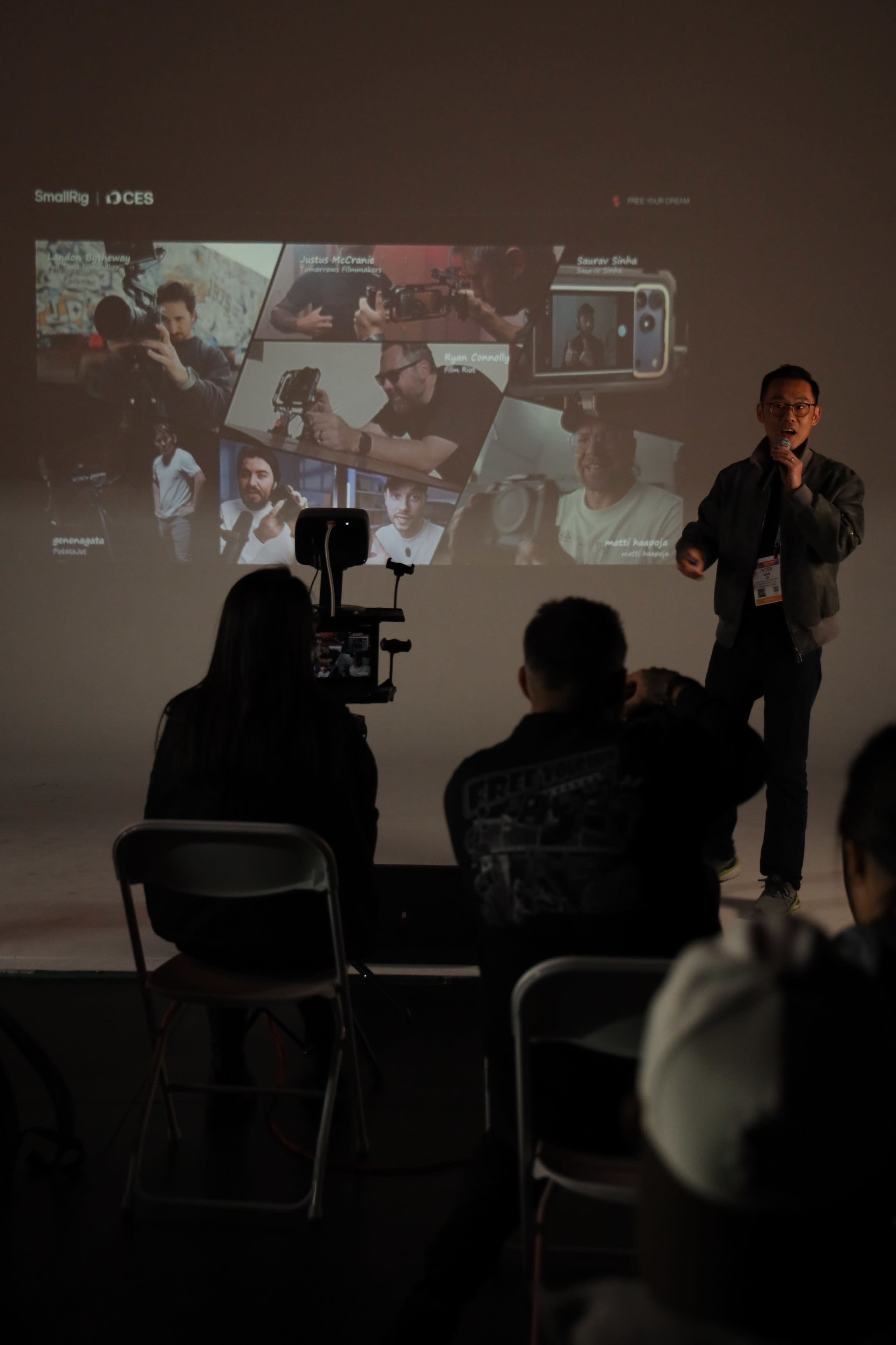 A presenter stands in front of an audience while displaying a slideshow featuring various camera equipment and filmmaking techniques.