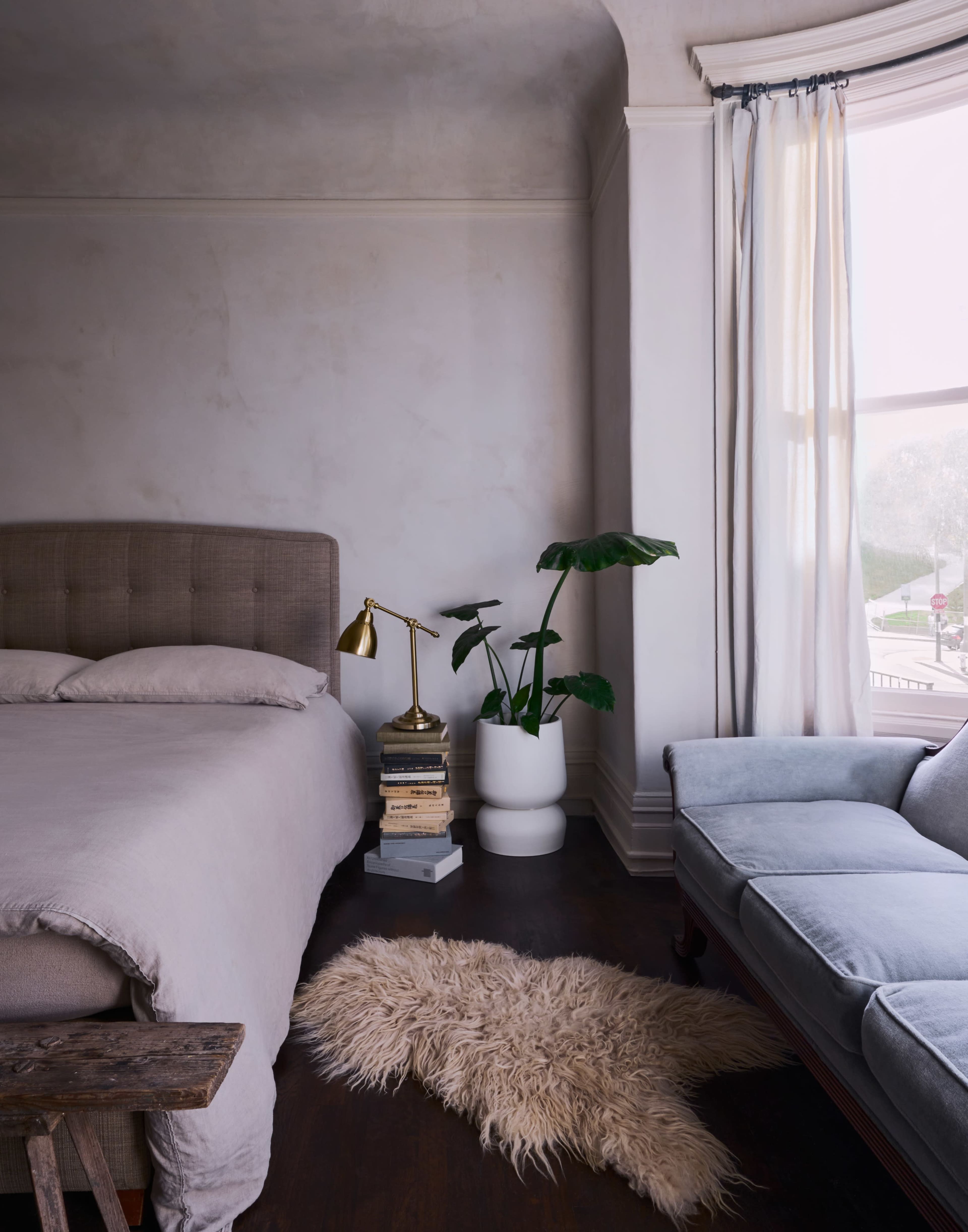 A bed with a beige headboard and bedding is positioned in a softly lit room alongside a reading chair, a lamp, a stack of books, and a potted plant near a large window.