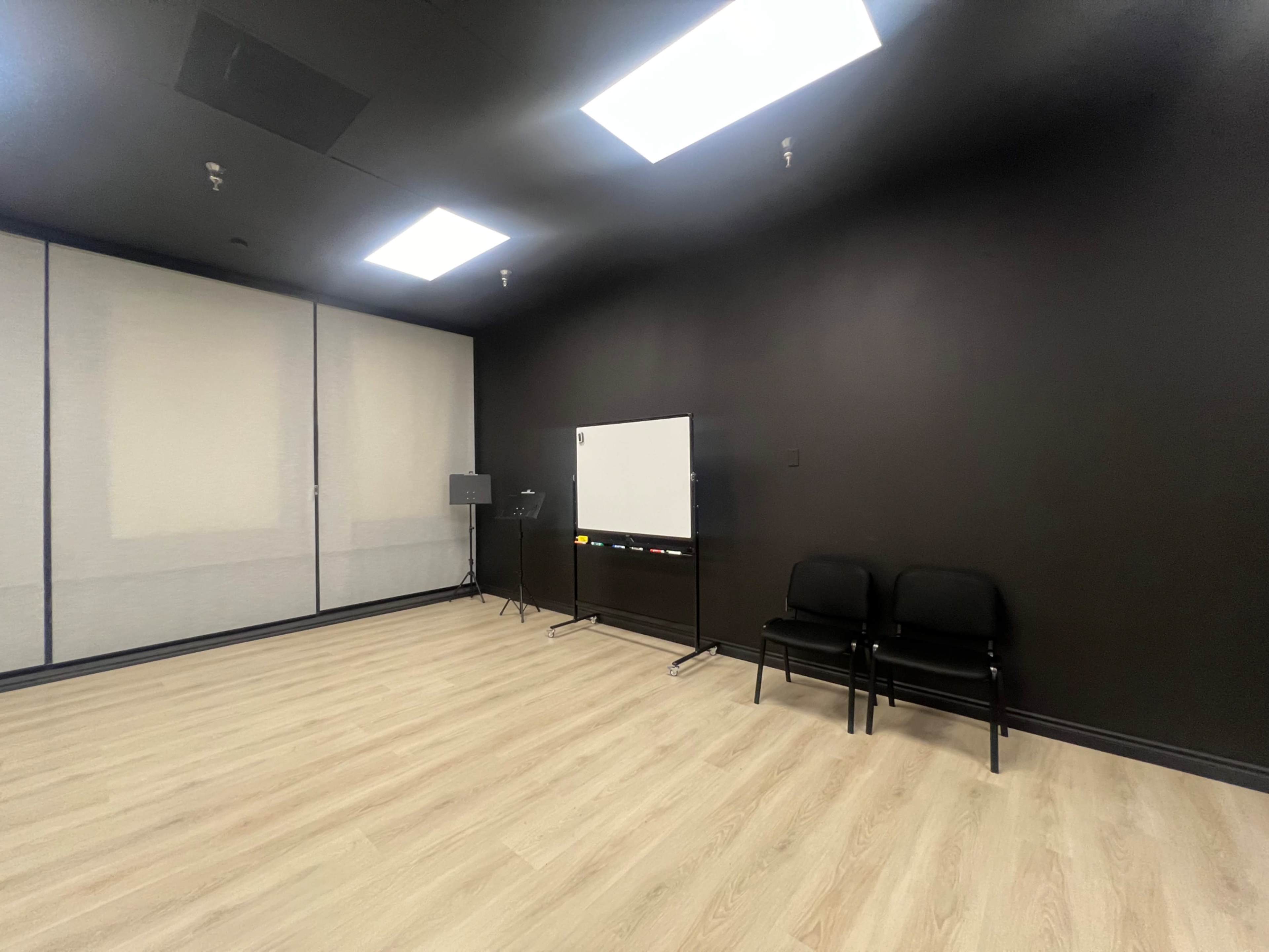 The image shows a minimally furnished room with two black chairs and a whiteboard on a stand against a dark wall.