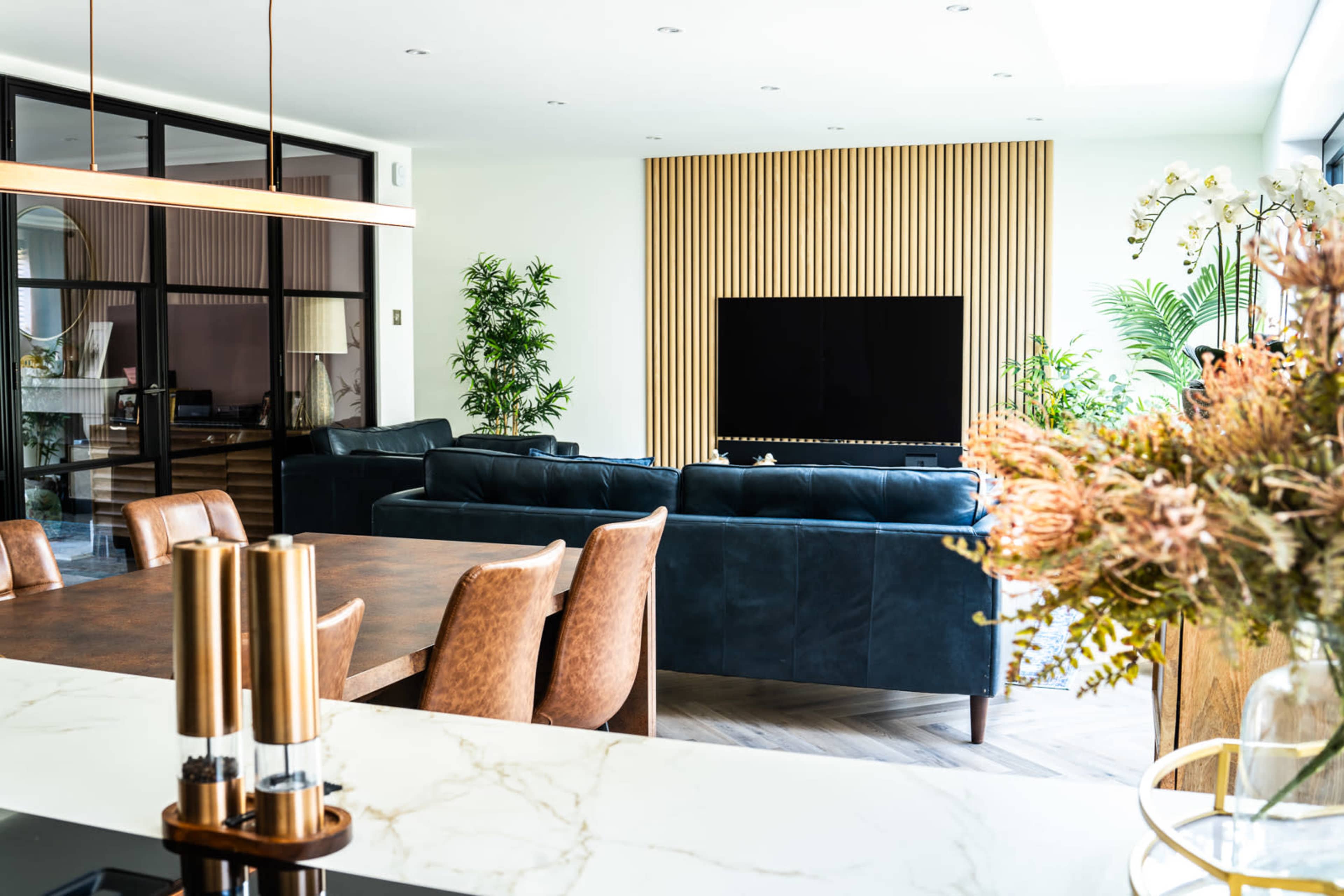 The image shows a modern living area with a dark blue sectional sofa, a wooden dining table, and a large television mounted on a wall with vertical wooden slats.