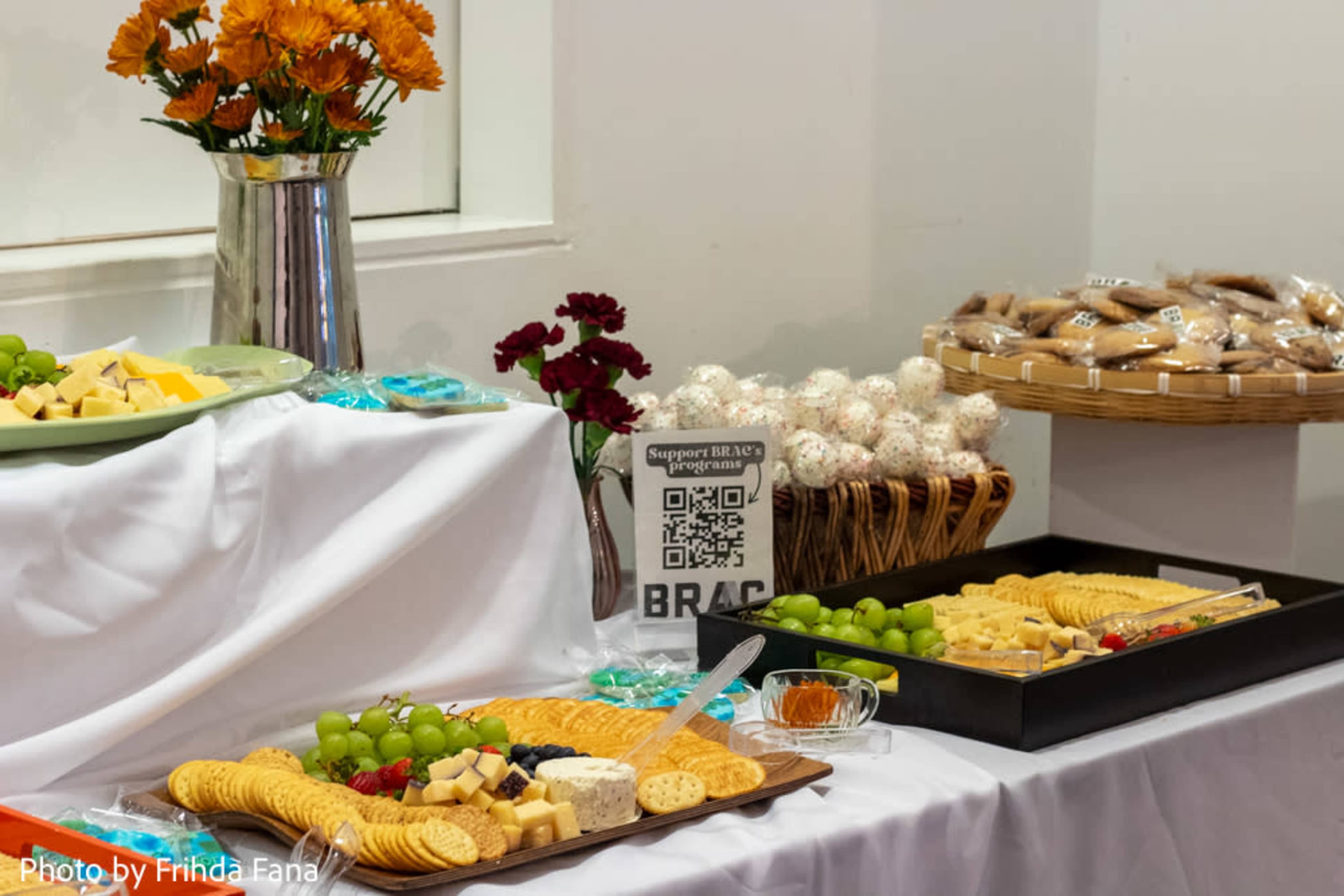 A table is set with an assortment of cheeses, crackers, grapes, and dessert items, accompanied by a QR code sign and flowers in a vase.