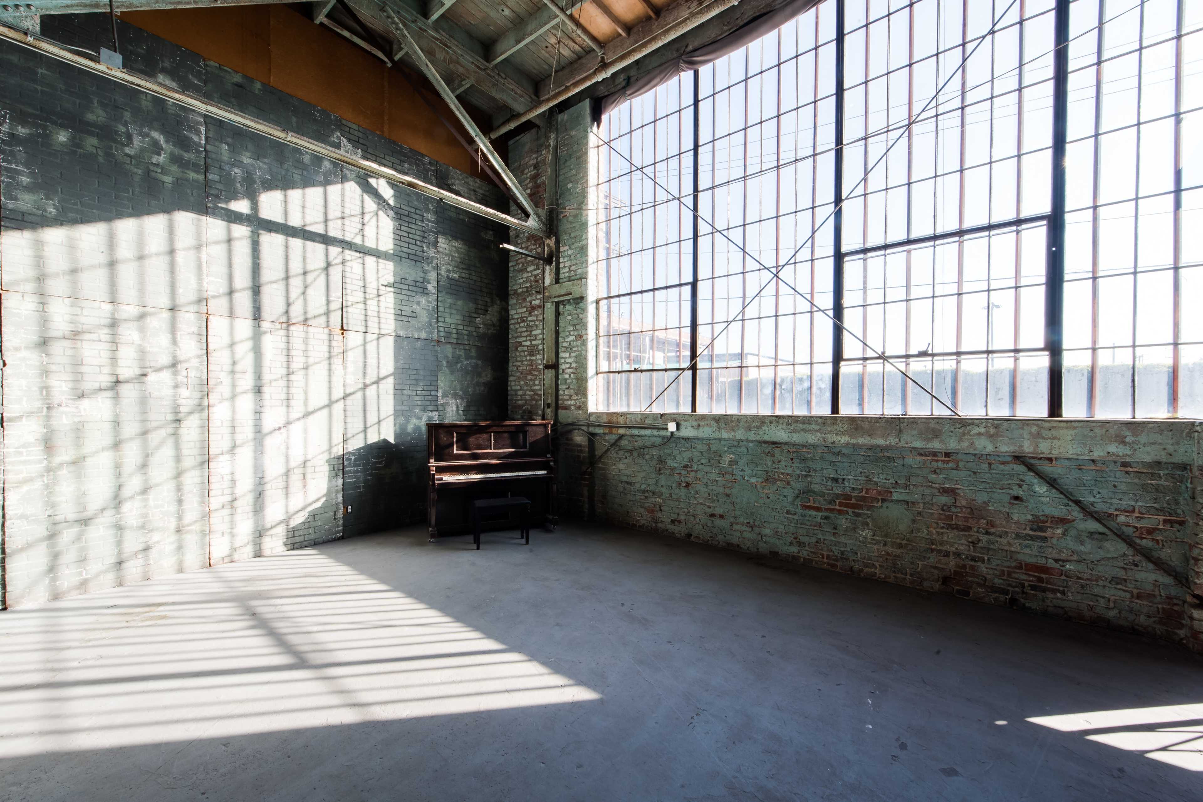 An empty industrial space with exposed brick walls, large windows, and a piano positioned against one wall.